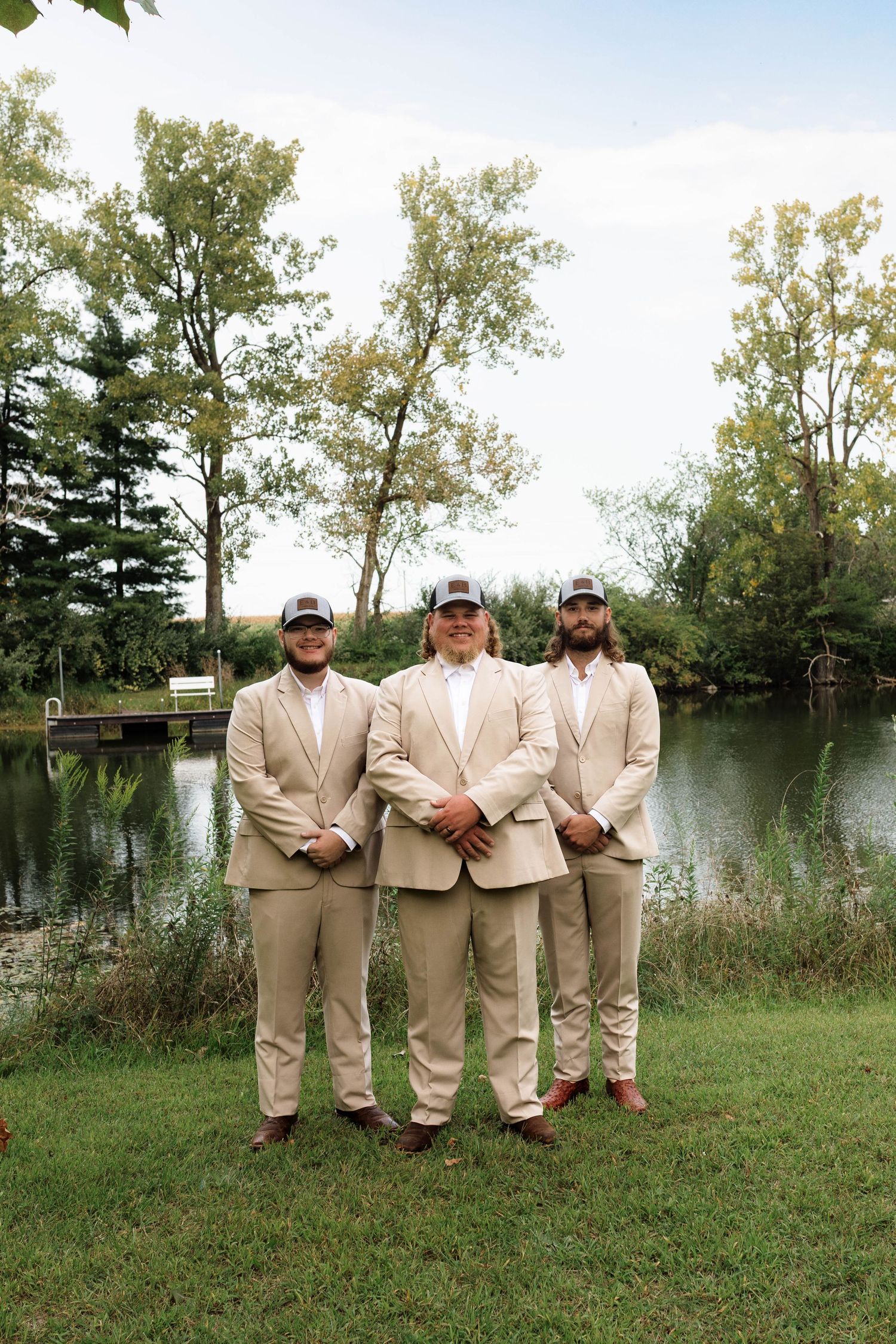 Three groomsmen in tan suits and trucker hats pose by a serene pond surrounded by lush green trees.