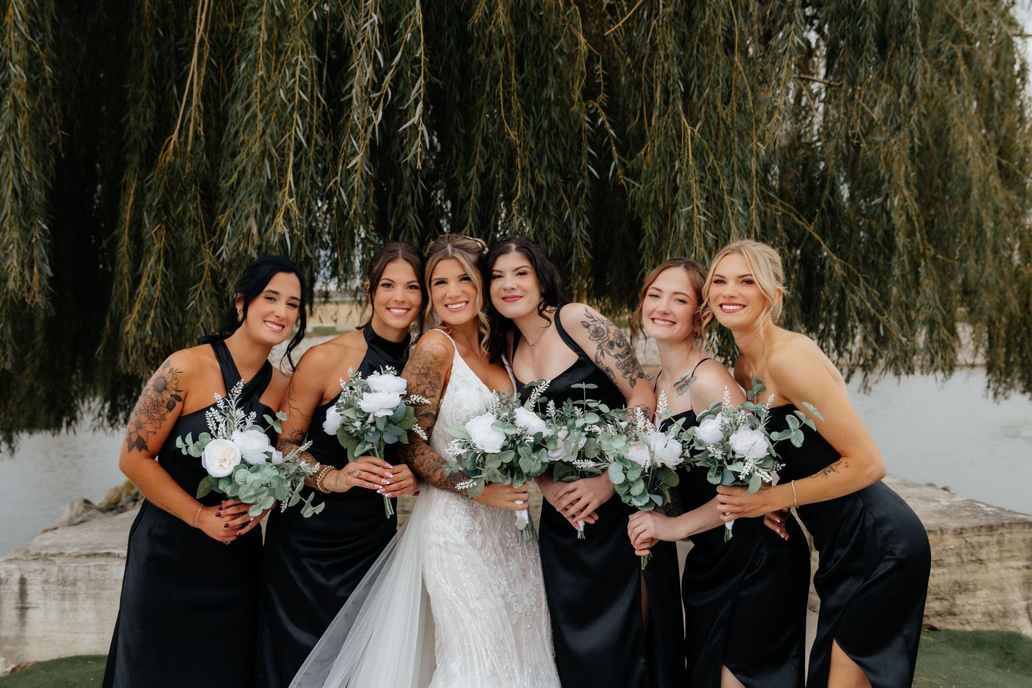 Bride with four bridesmaids in black dresses holding white bouquets under willow tree.