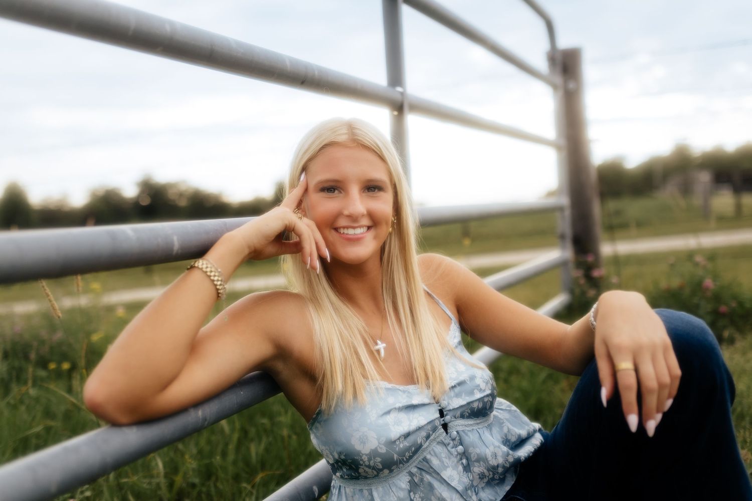 A smiling blonde girl leans against a metal gate in a green field wearing a denim top and jeans.