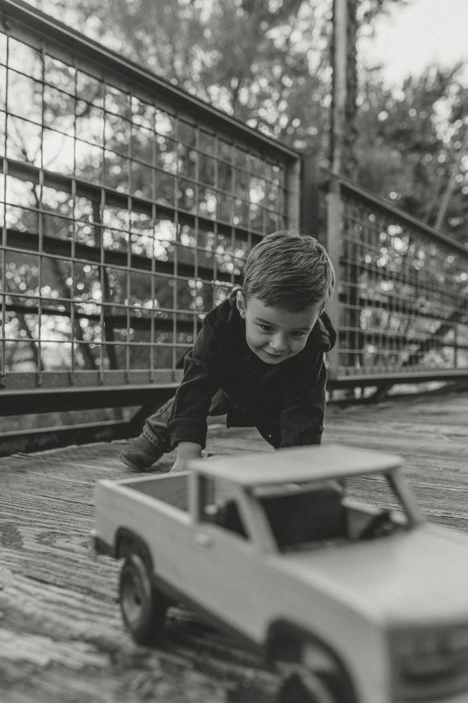 Black and white photo of young boy playing with toy pickup truck on wooden bridge.
