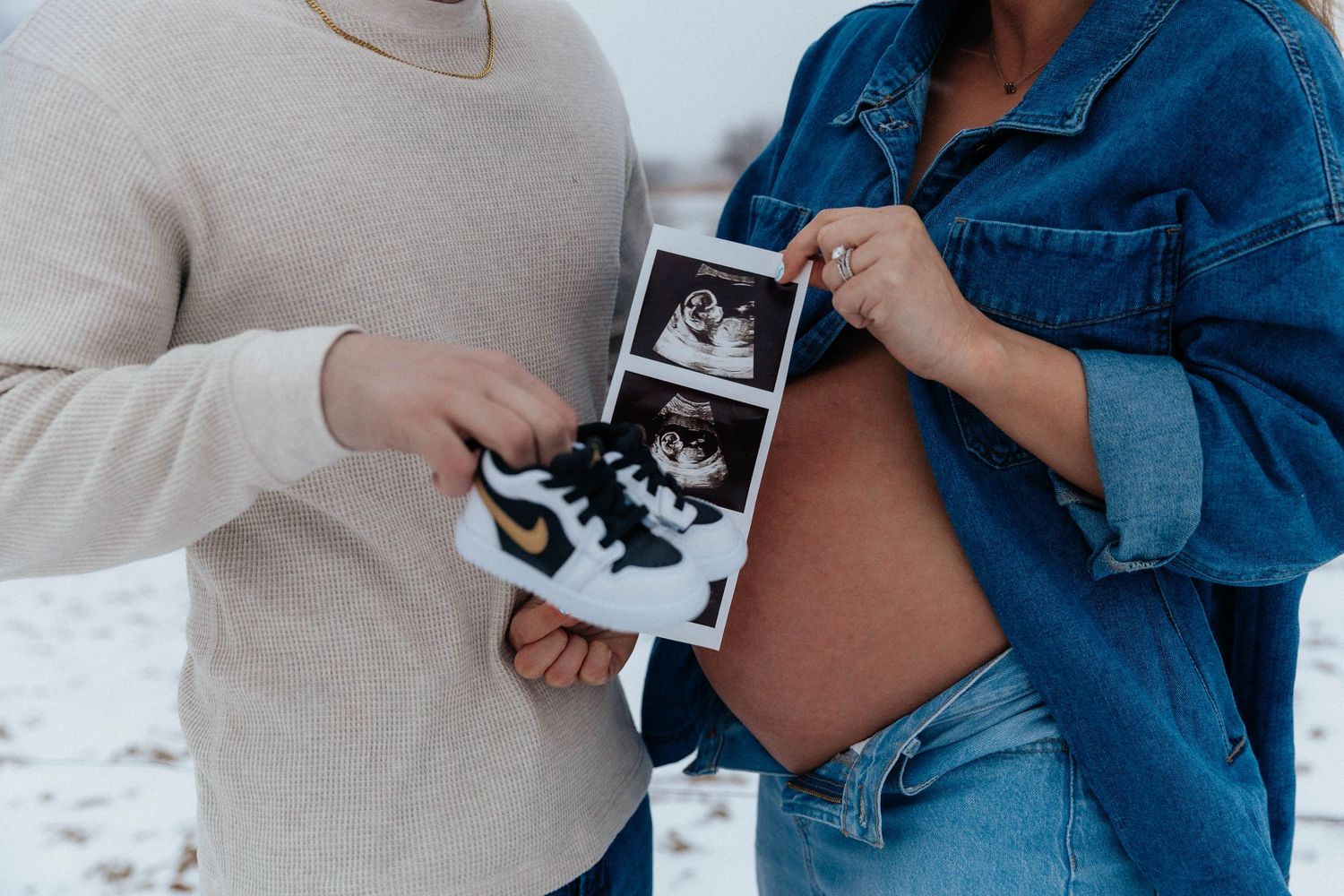 Couple holding baby shoes and ultrasound photo during snowy outdoor maternity shoot.