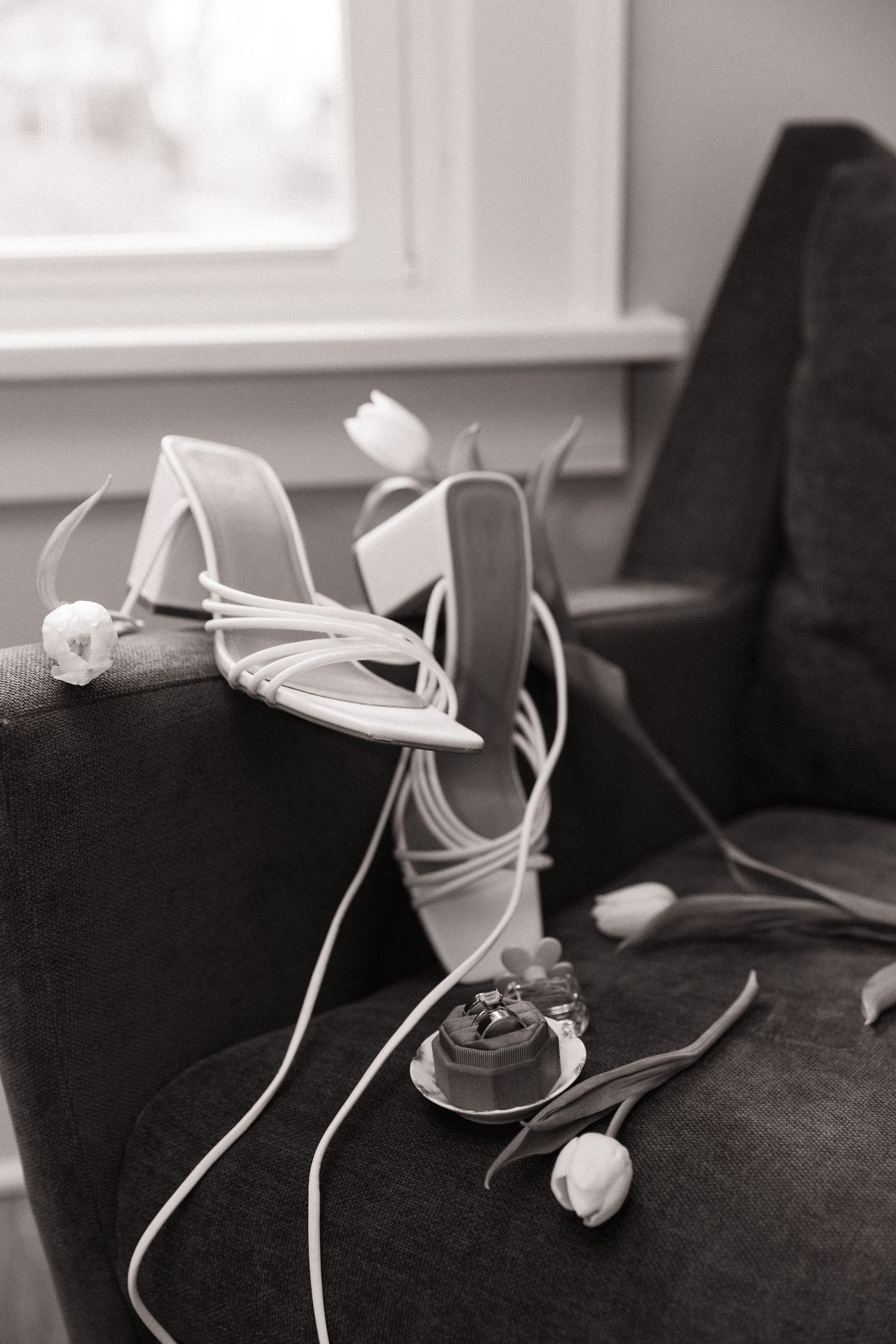 Black and white photo of strappy bridal heels and ring box with tulips on velvet chair.