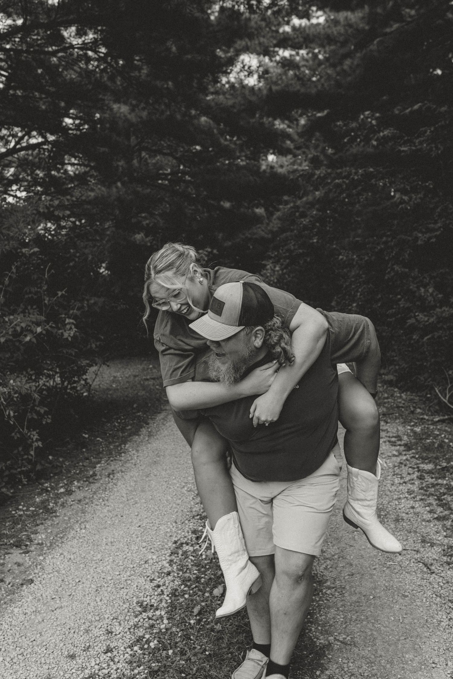 Black and white photo of groom giving bride a piggyback ride on a gravel path.