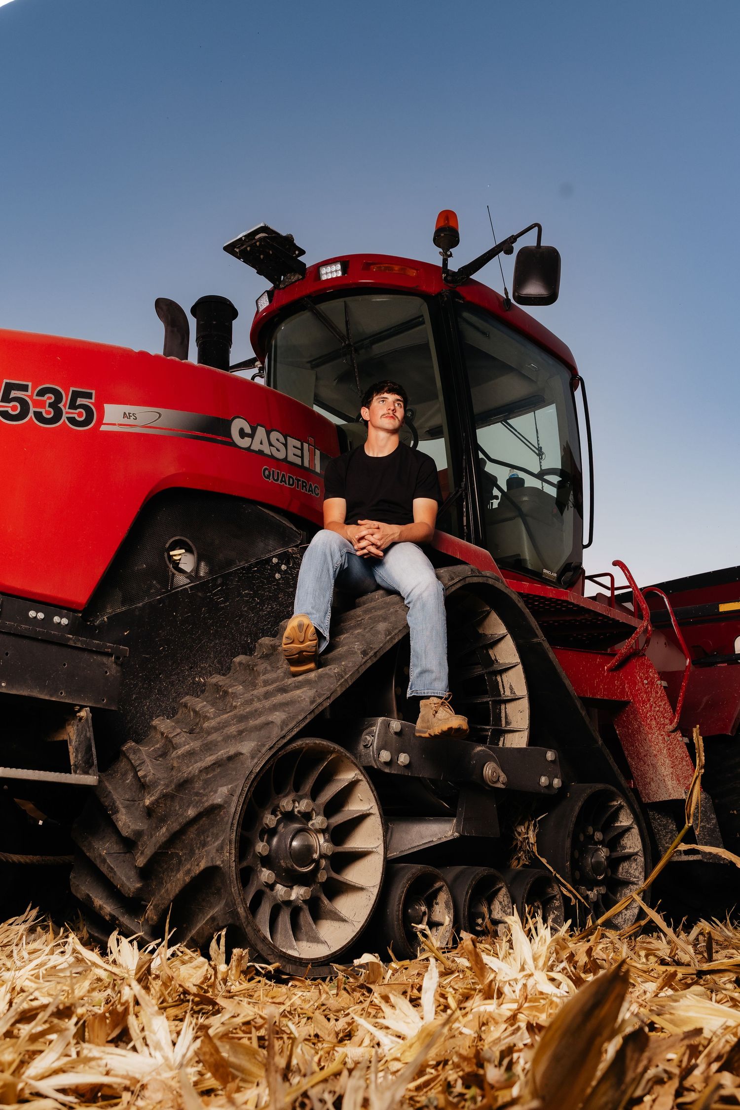 Young man sits on red Case IH 535 tracked tractor in a harvested corn field at dusk.