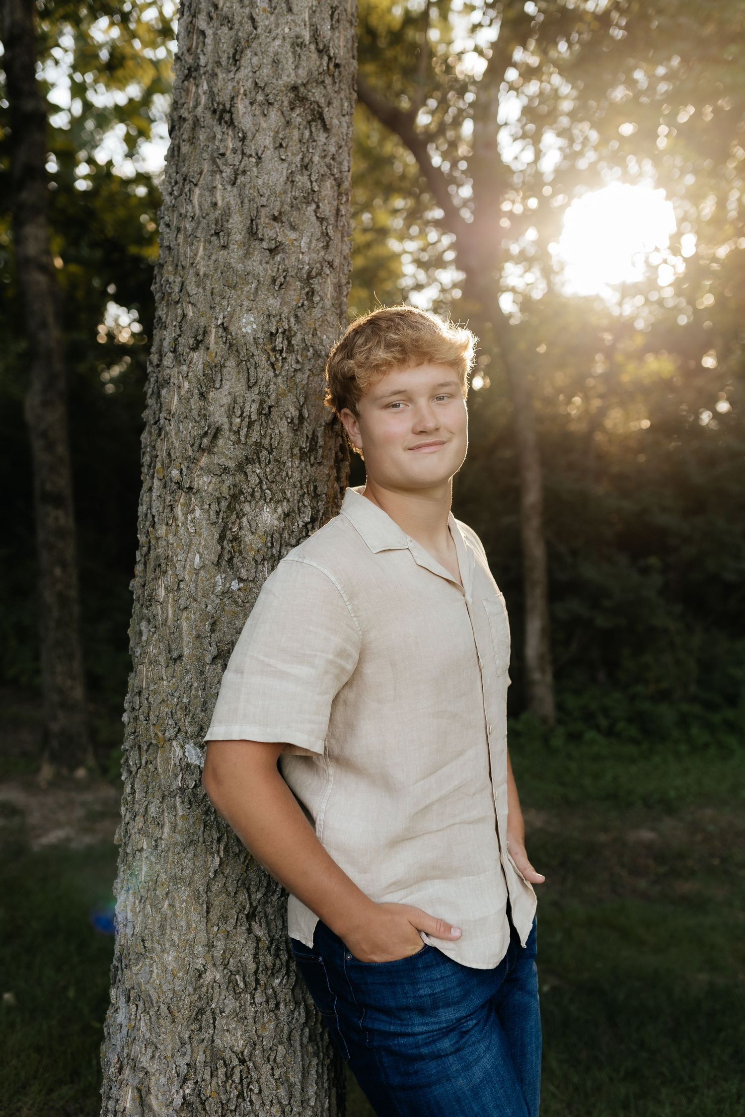 Young man in a beige short-sleeve shirt and jeans leaning against a tree trunk at sunset.