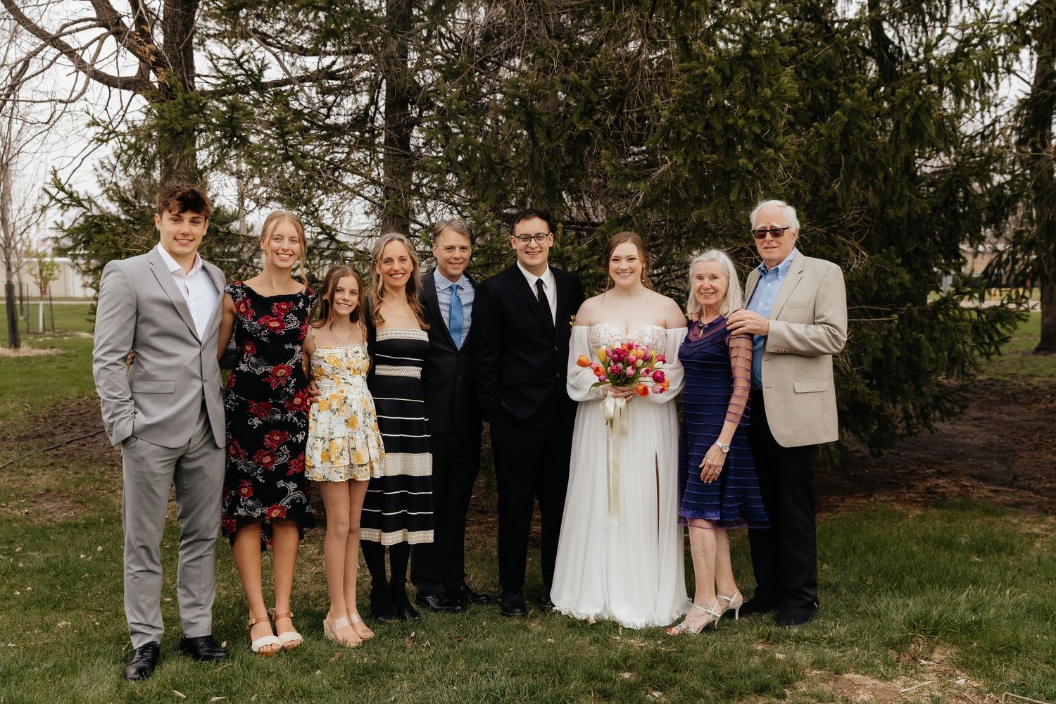 Wedding party poses outdoors with bride holding colorful bouquet surrounded by family in formal attire near evergreen trees.