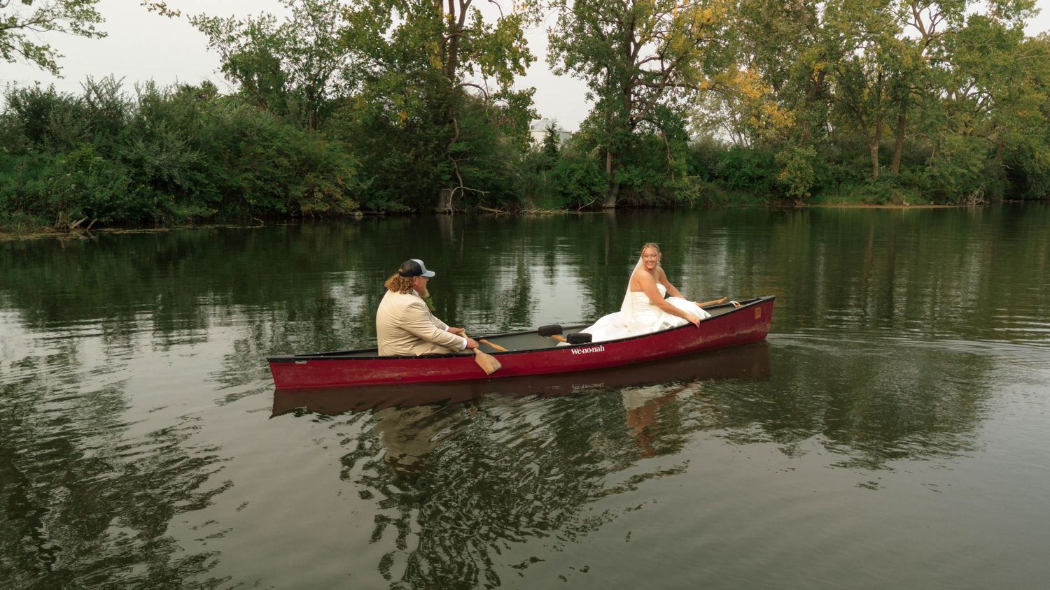 Bride and groom rowing a red canoe on a serene tree-lined river.