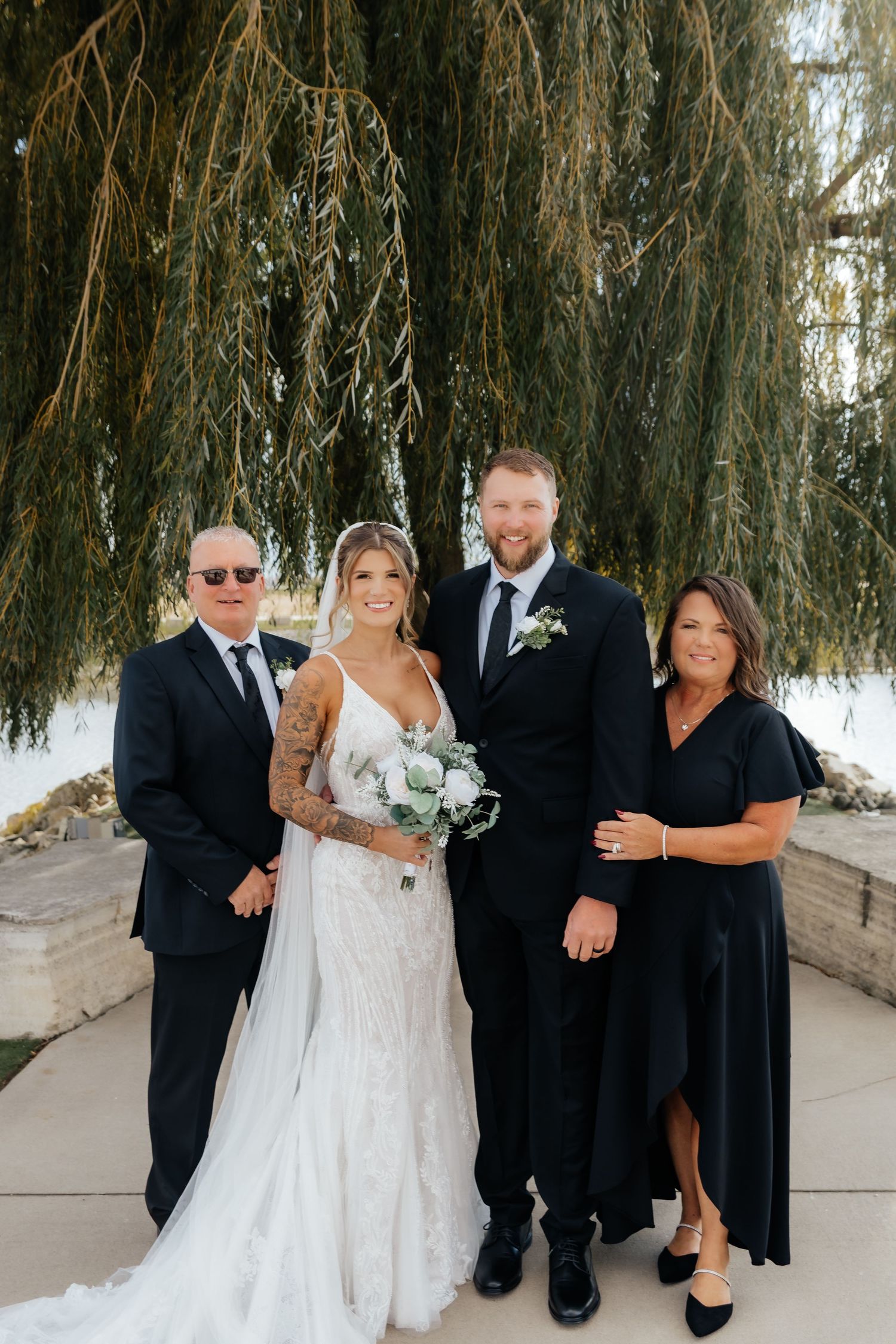 Wedding couple poses with two family members under weeping willow tree at outdoor venue.