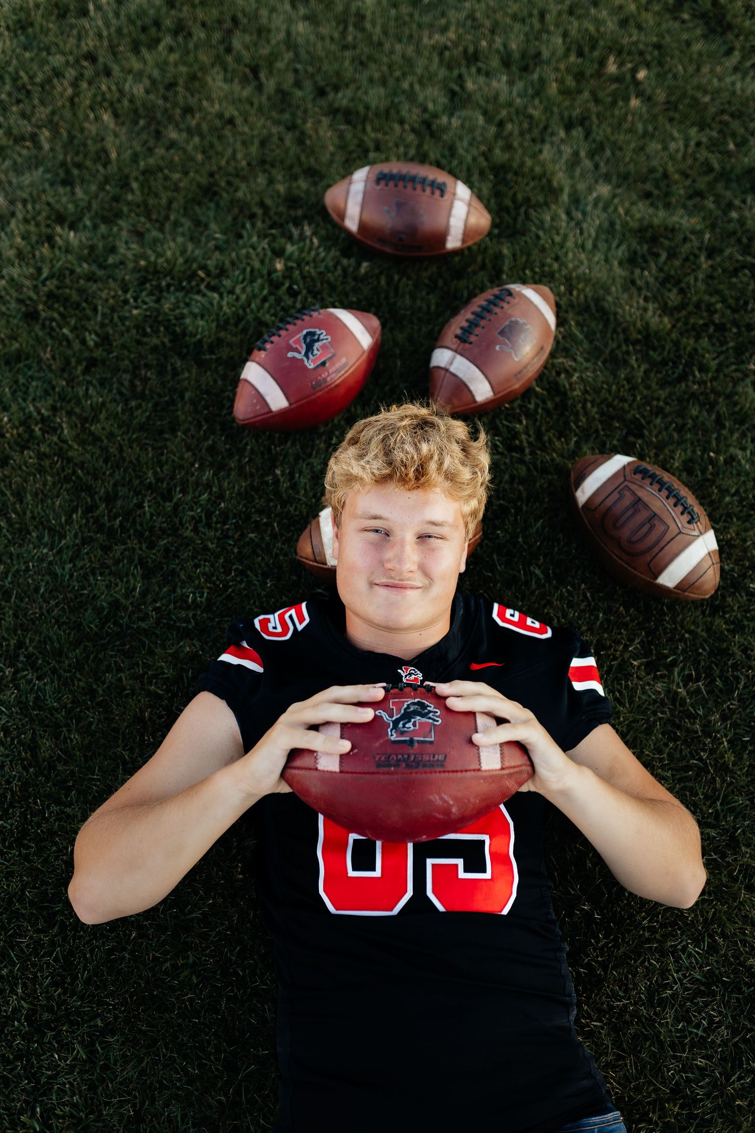 Young football player in black jersey holding a football lying on grass surrounded by footballs.