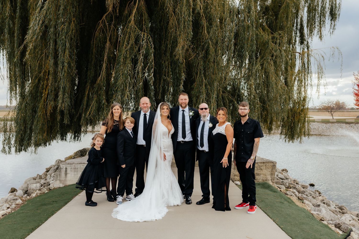 Bride and groom pose with large family group near waterfront under sweeping willow tree branches.