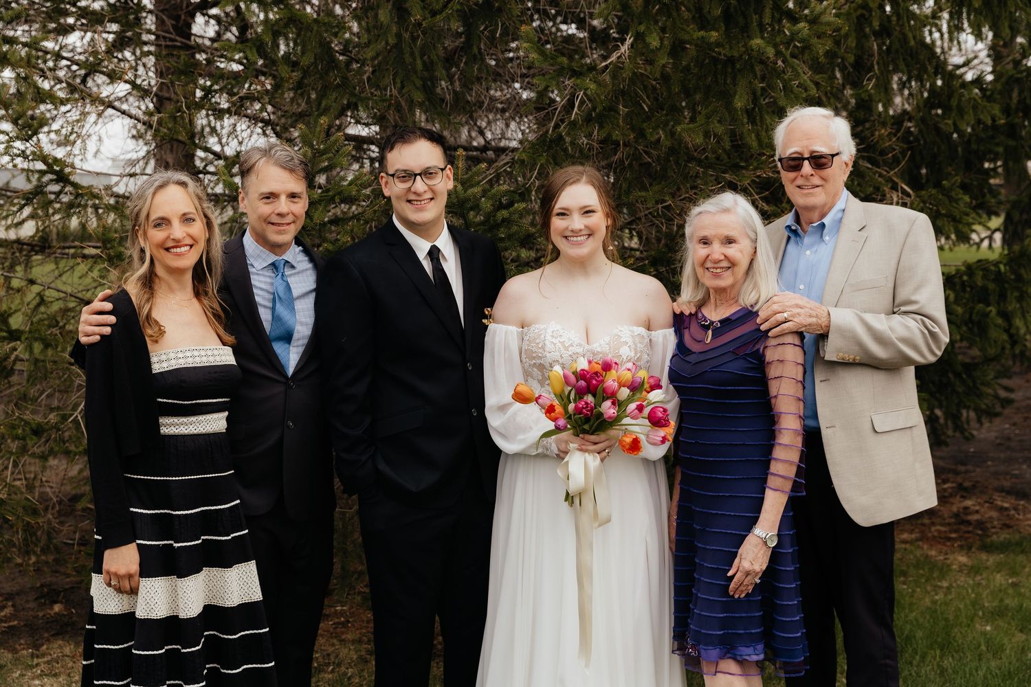 Bride with colorful bouquet poses with groom and four family members outdoors near green trees on wedding day.
