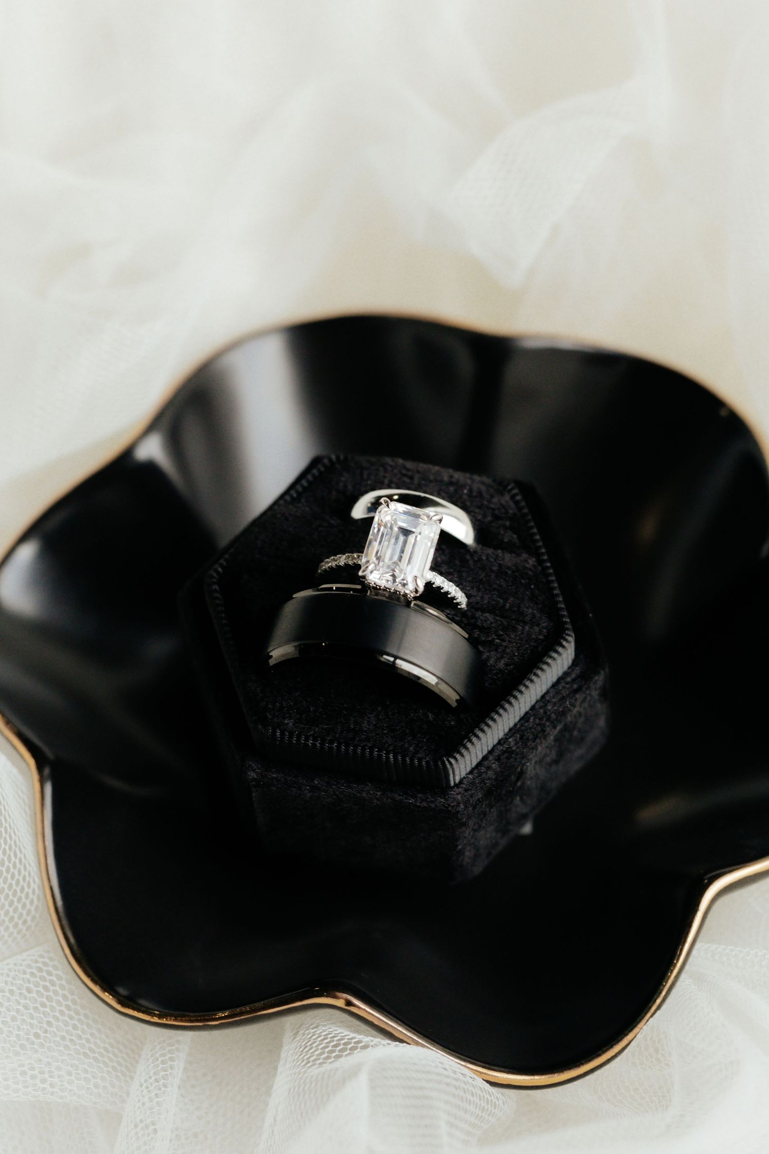 Emerald cut diamond engagement ring and black wedding band displayed in black velvet hexagon ring box.