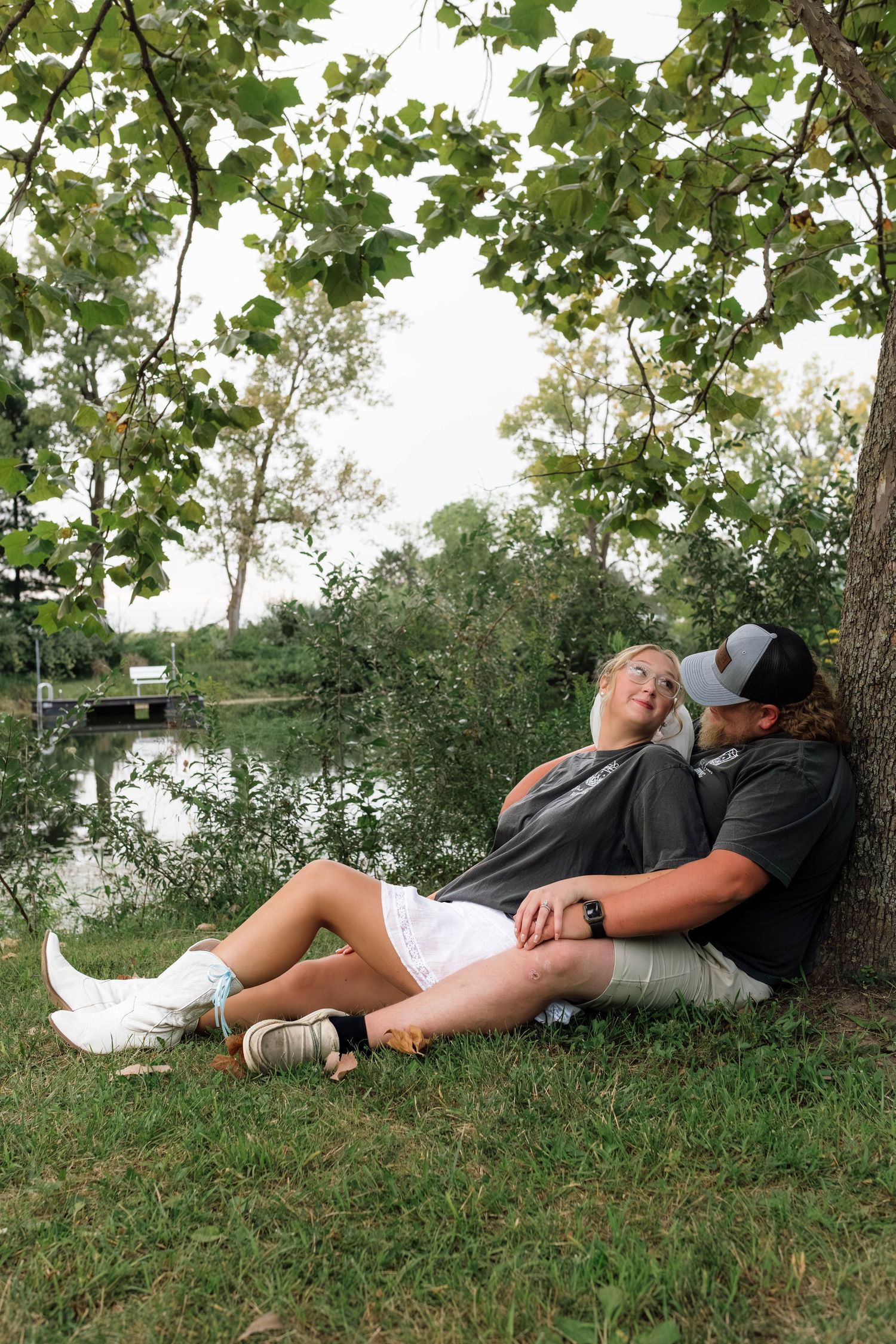 Newlywed couple relaxing against a tree by a peaceful lake in casual wedding attire.