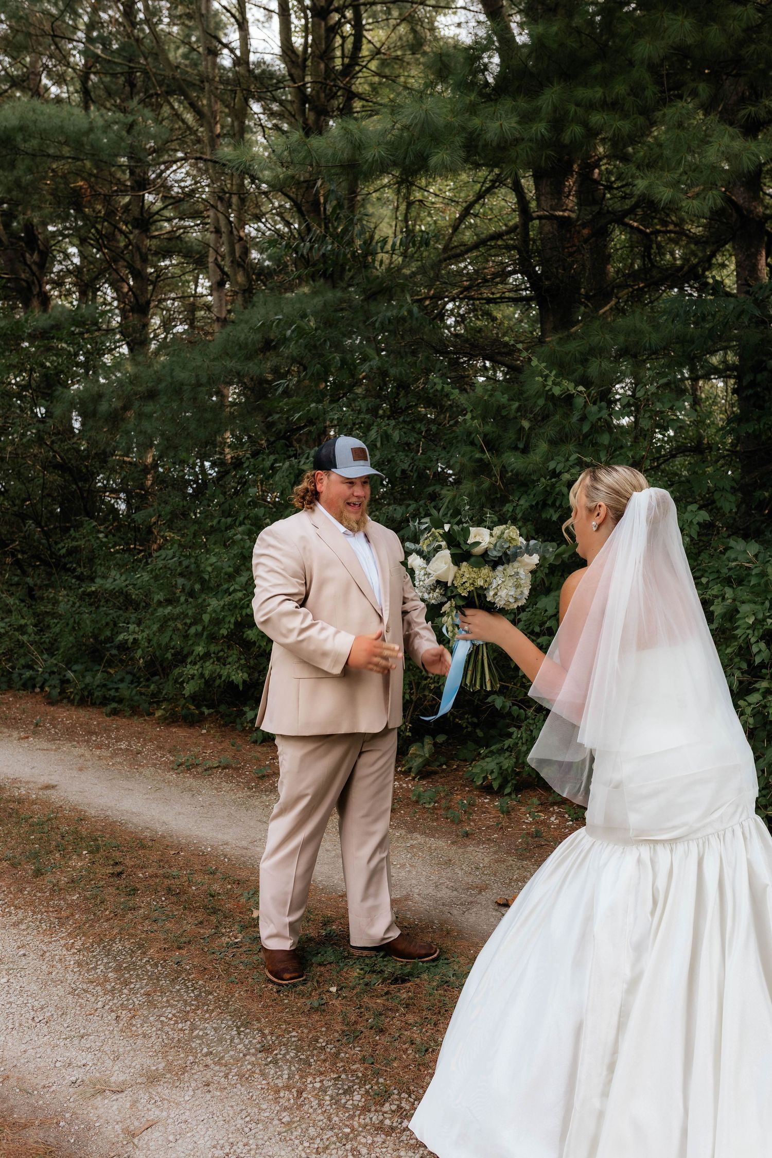 Groom in beige suit and cap reacts with joy seeing bride in white gown holding bouquet on wooded path.