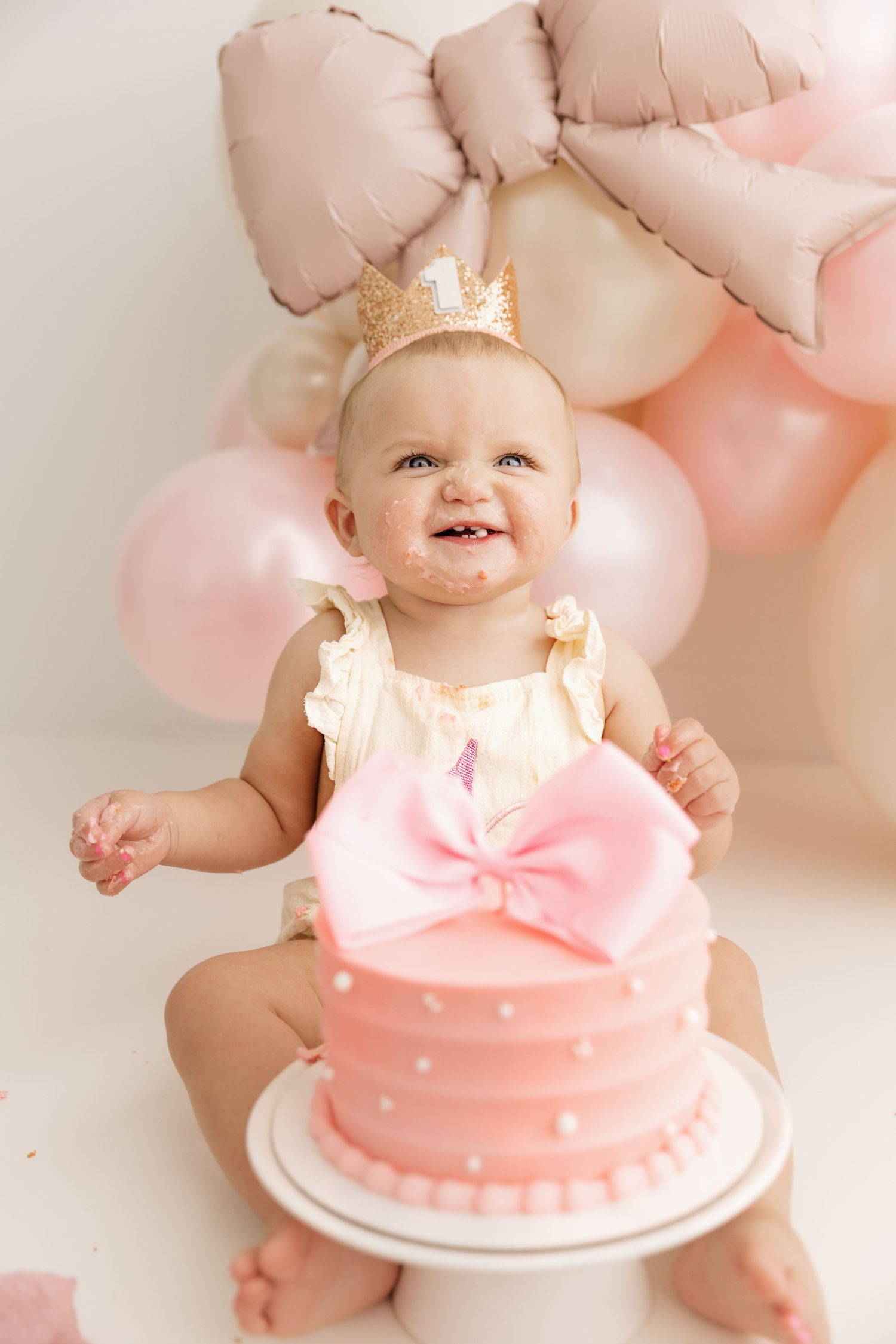 Joyful baby girl with cake-smeared face wearing gold crown sitting with pink birthday cake and balloons.