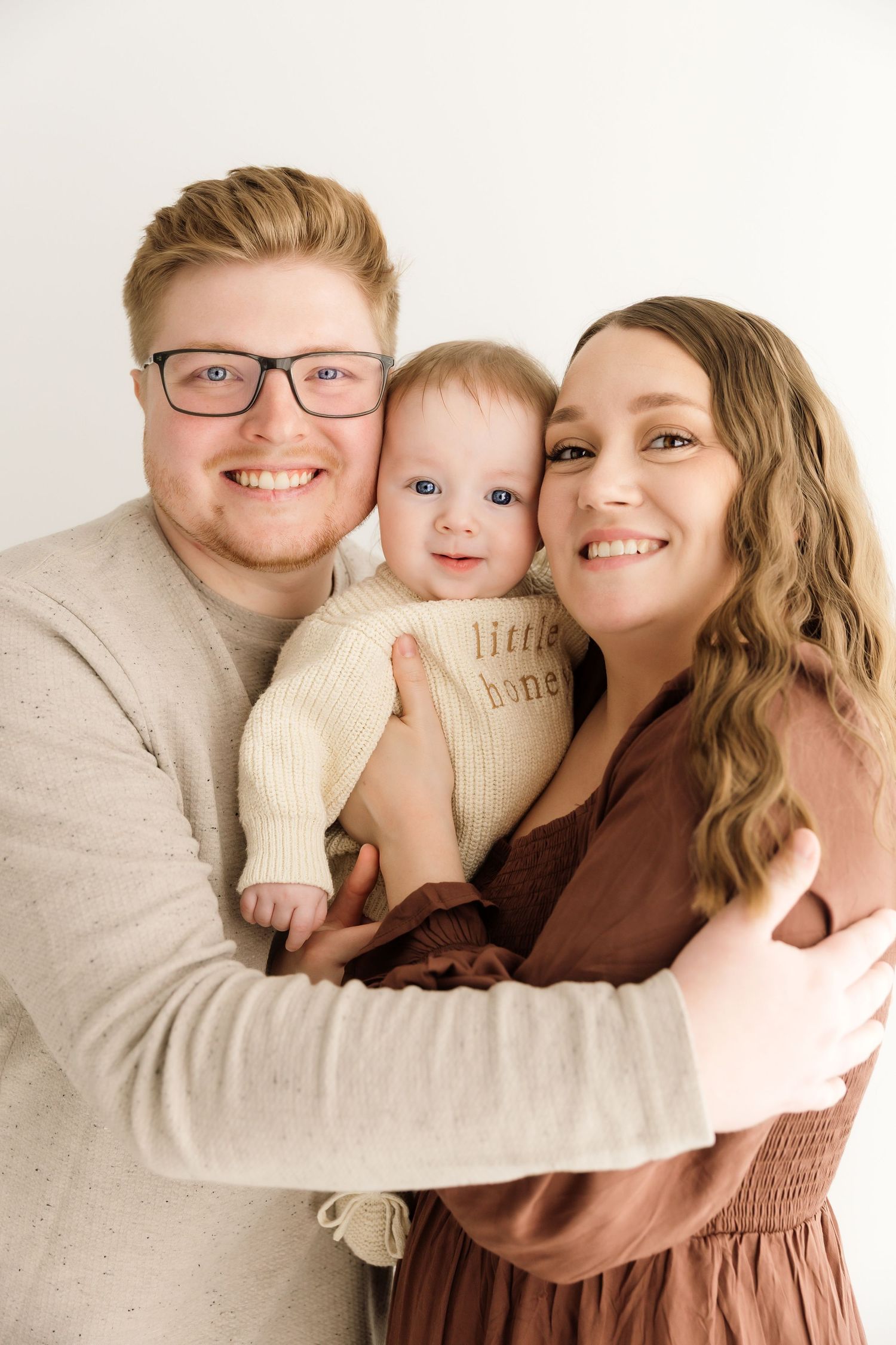 Happy young family with smiling parents holding their baby dressed in cream knit 'little honey' outfit.