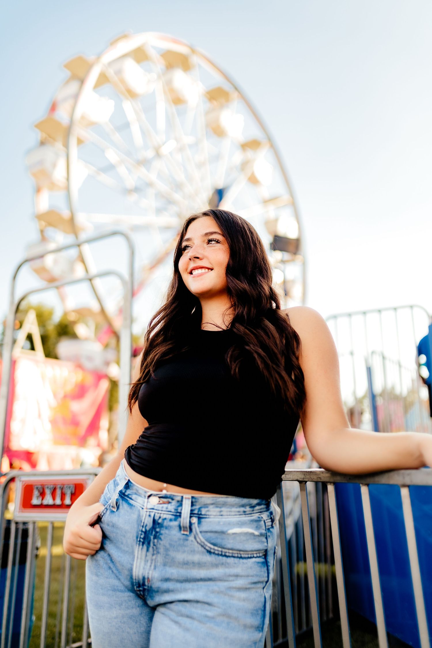 Smiling young woman in black crop top and jeans leaning on railing near ferris wheel.