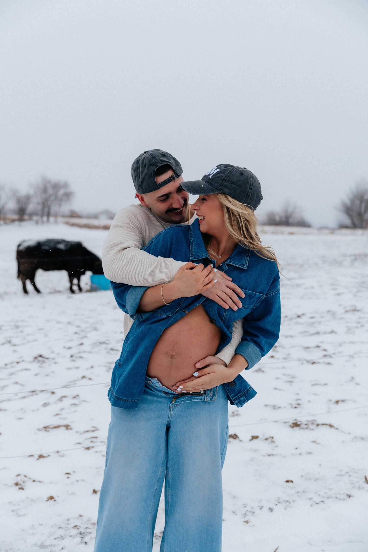 Couple embraces showing pregnant belly in snowy farm field with black cow in background.