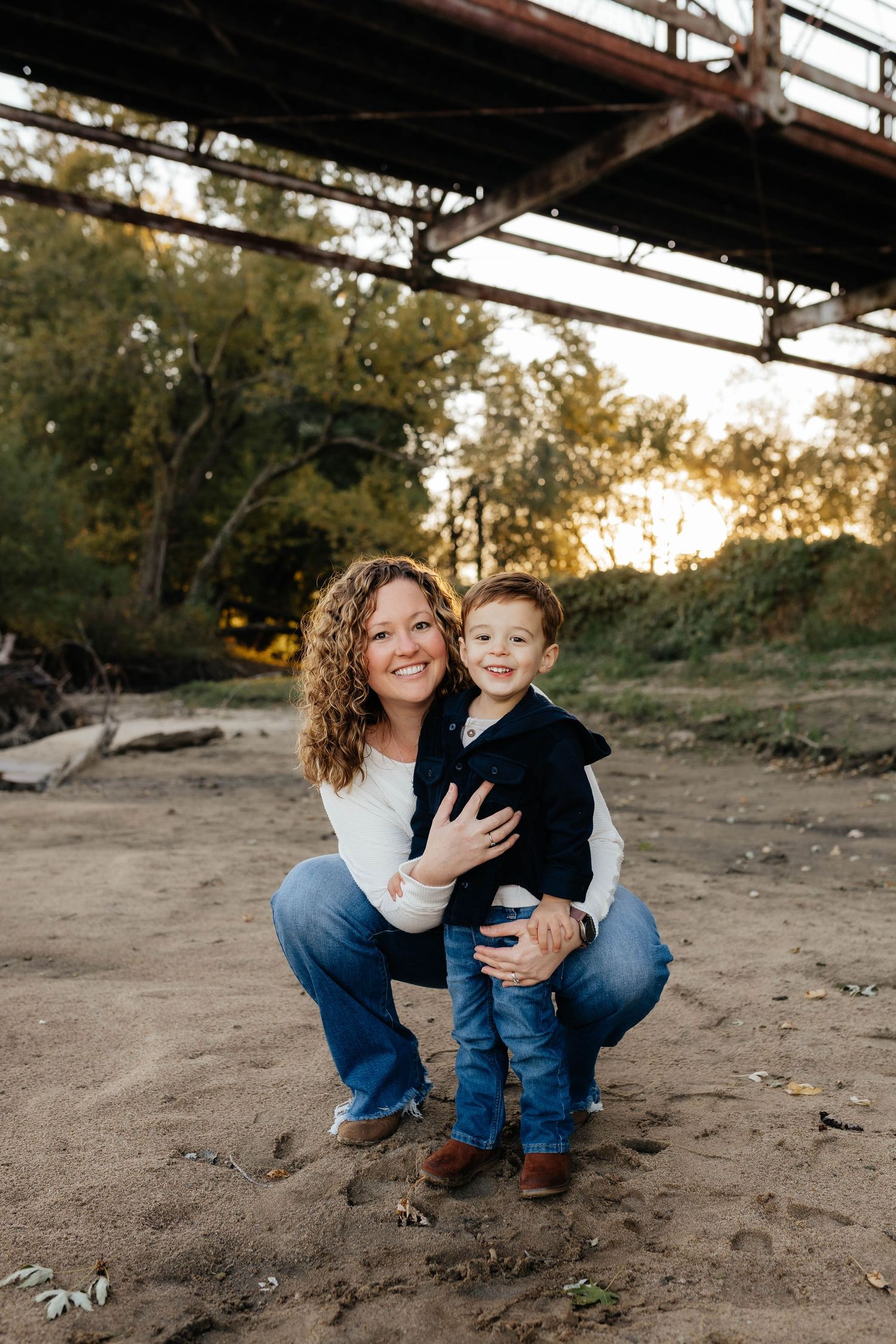 Smiling mother crouches with young toddler son outdoors near rustic bridge at golden hour.