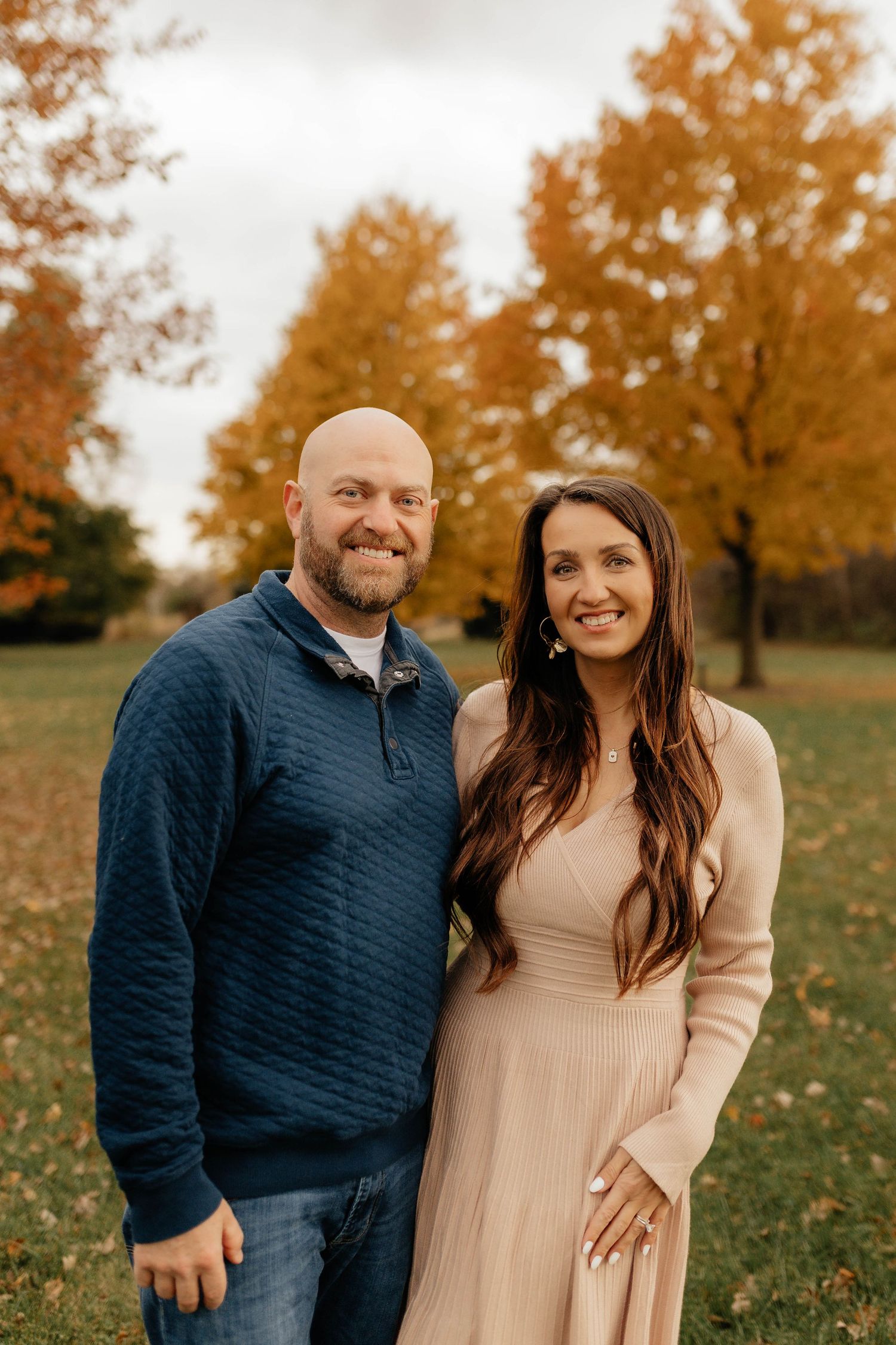 Smiling couple posing outdoors in autumn park with colorful orange foliage, man in navy sweater and woman in beige dress.