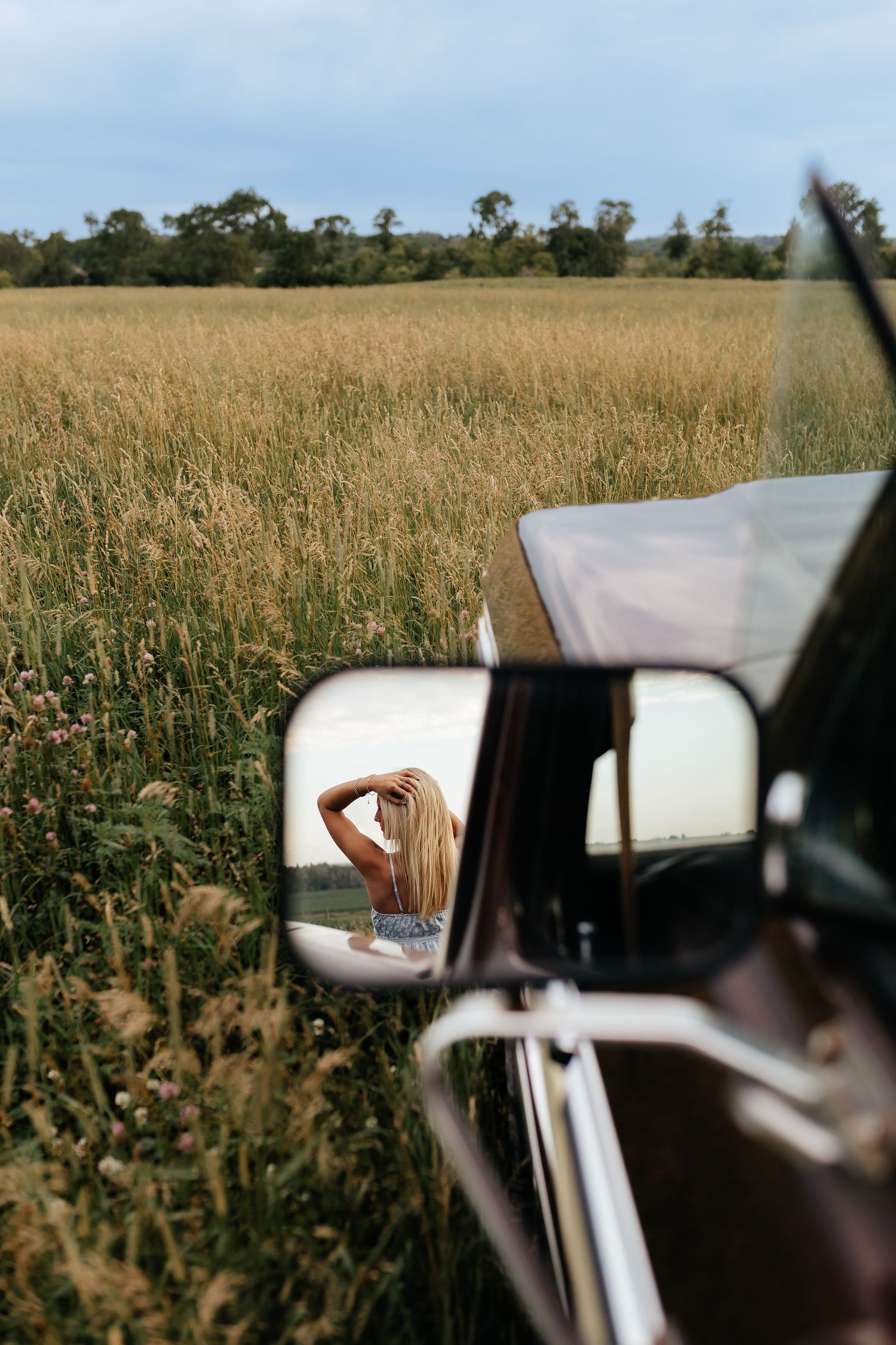 A blonde woman standing in a tall grass field is reflected in the side mirror of a vintage truck.