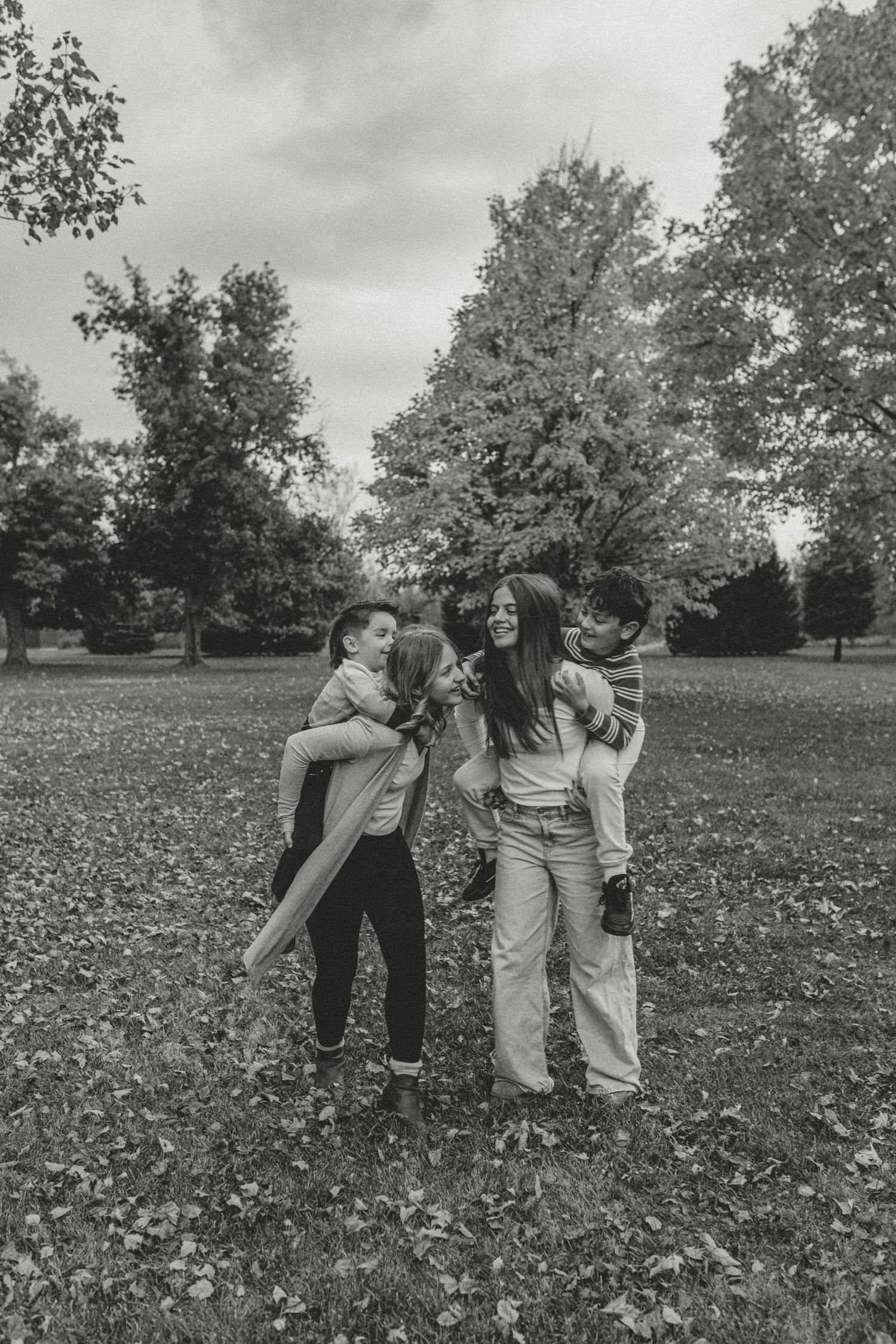 Black and white photo of a joyful family playing together in a park surrounded by trees on a cloudy day.