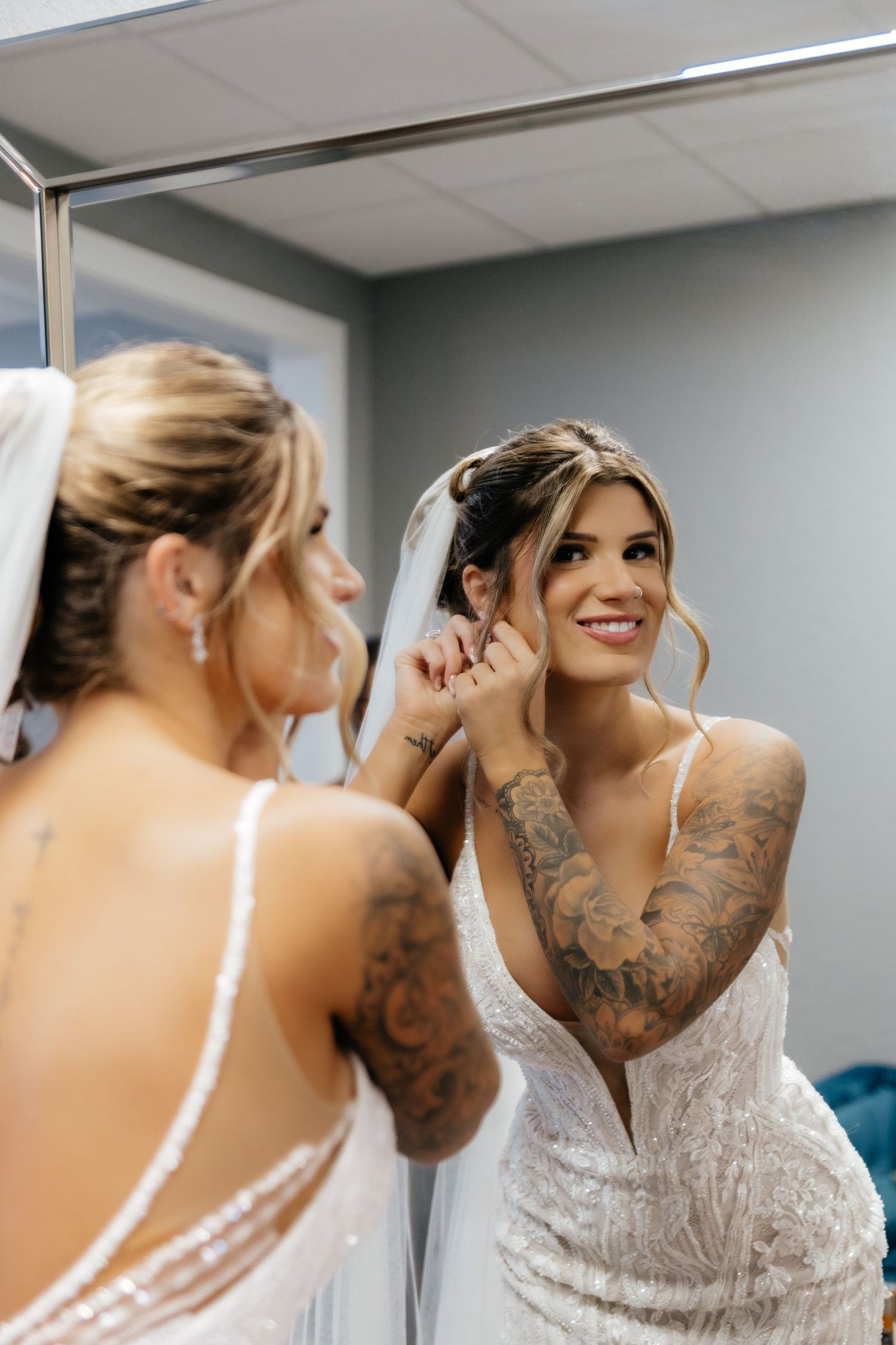 Tattooed bride in lace wedding gown smiling while putting on earrings in a mirror.