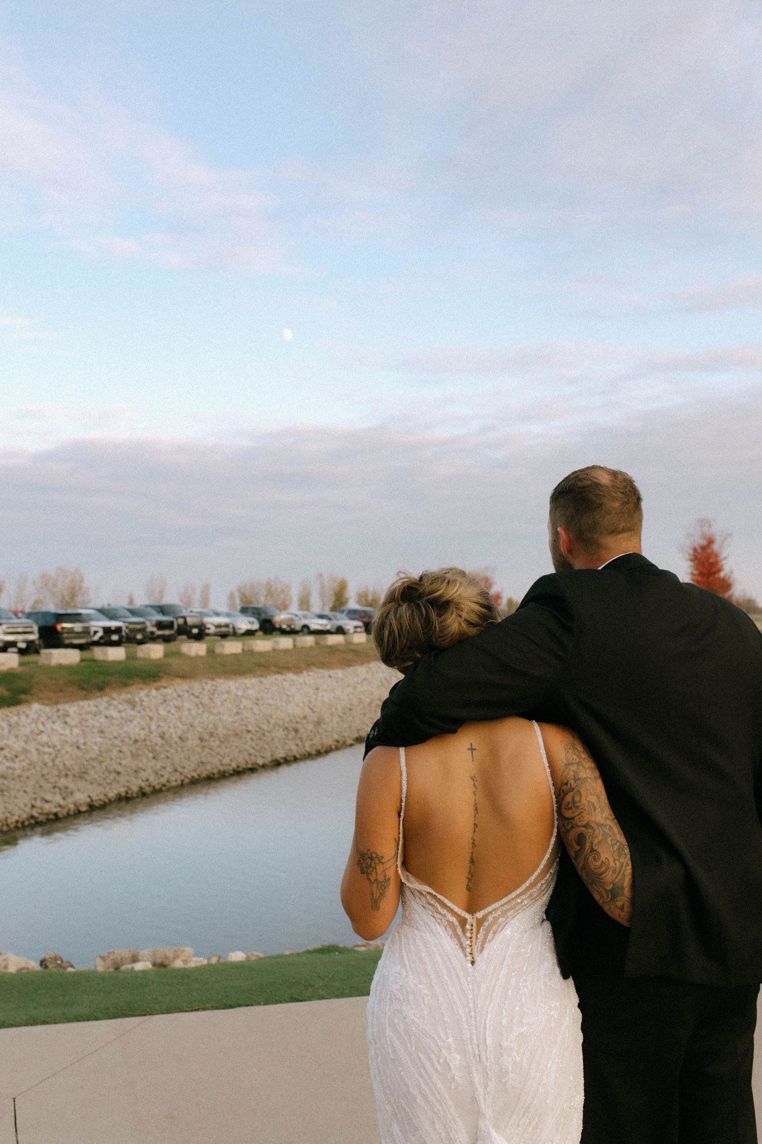 Tattooed bride and groom embrace outdoors at dusk overlooking a scenic waterway after wedding.