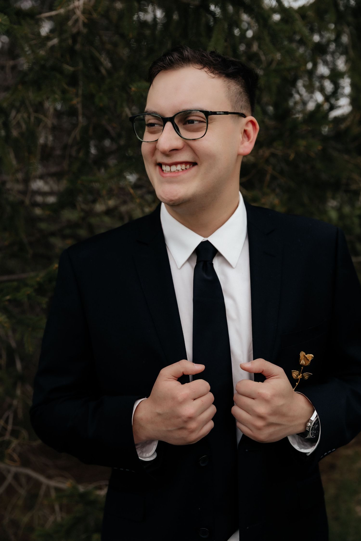 Smiling groom in black suit and glasses adjusts jacket lapels outdoors near evergreen trees.