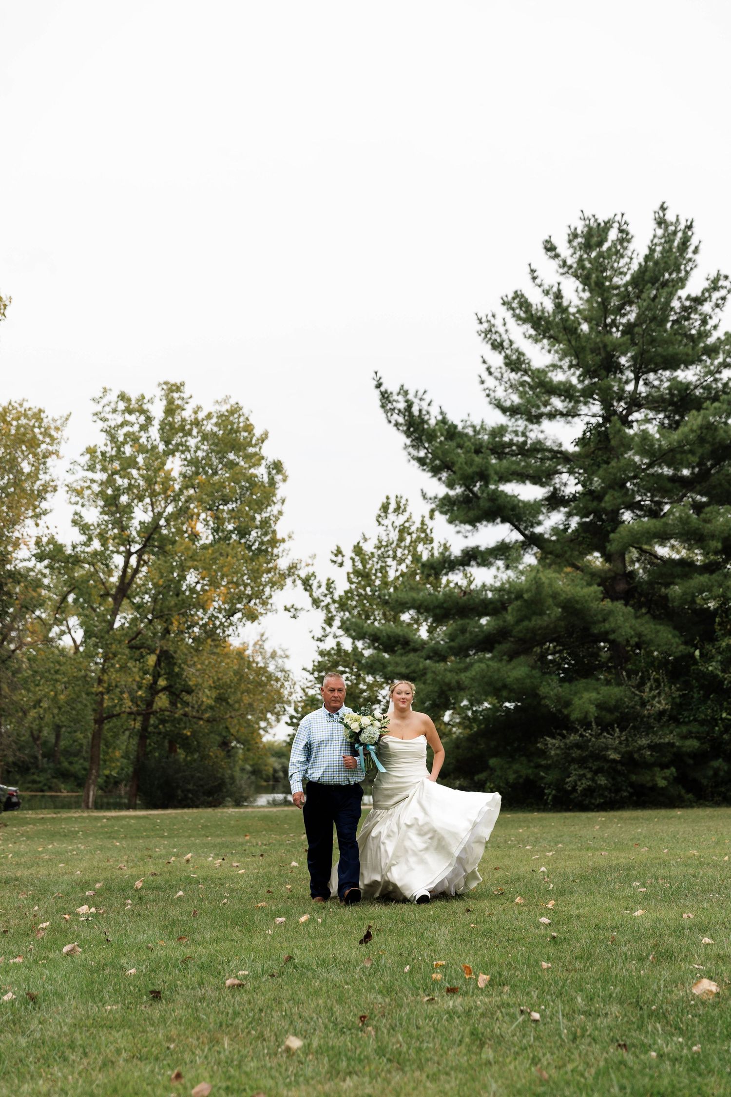 Bride in white gown walks with escort across green lawn at outdoor fall wedding ceremony.