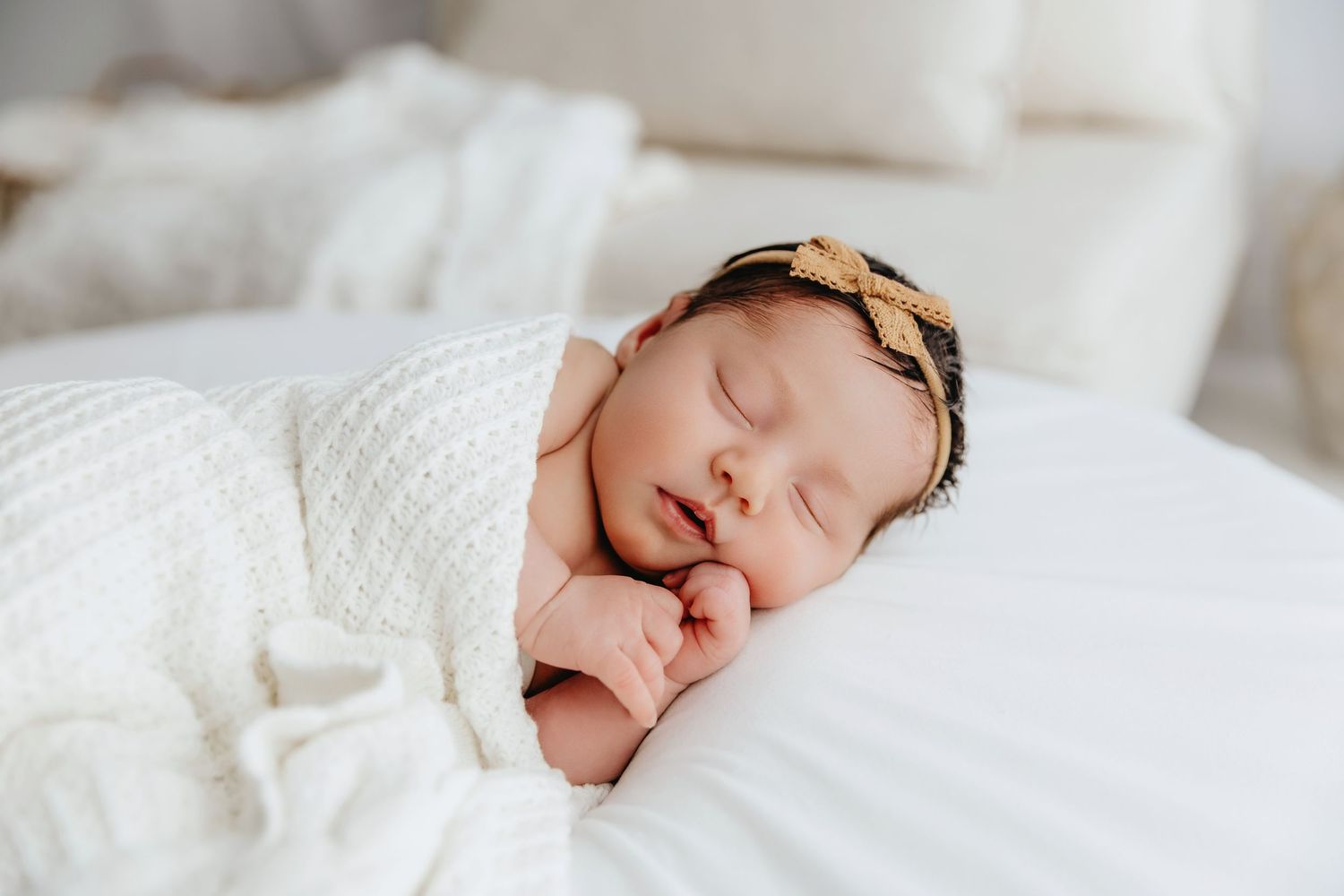 Newborn baby sleeping peacefully wrapped in white knit swaddle with gold bow headband on white bedding.