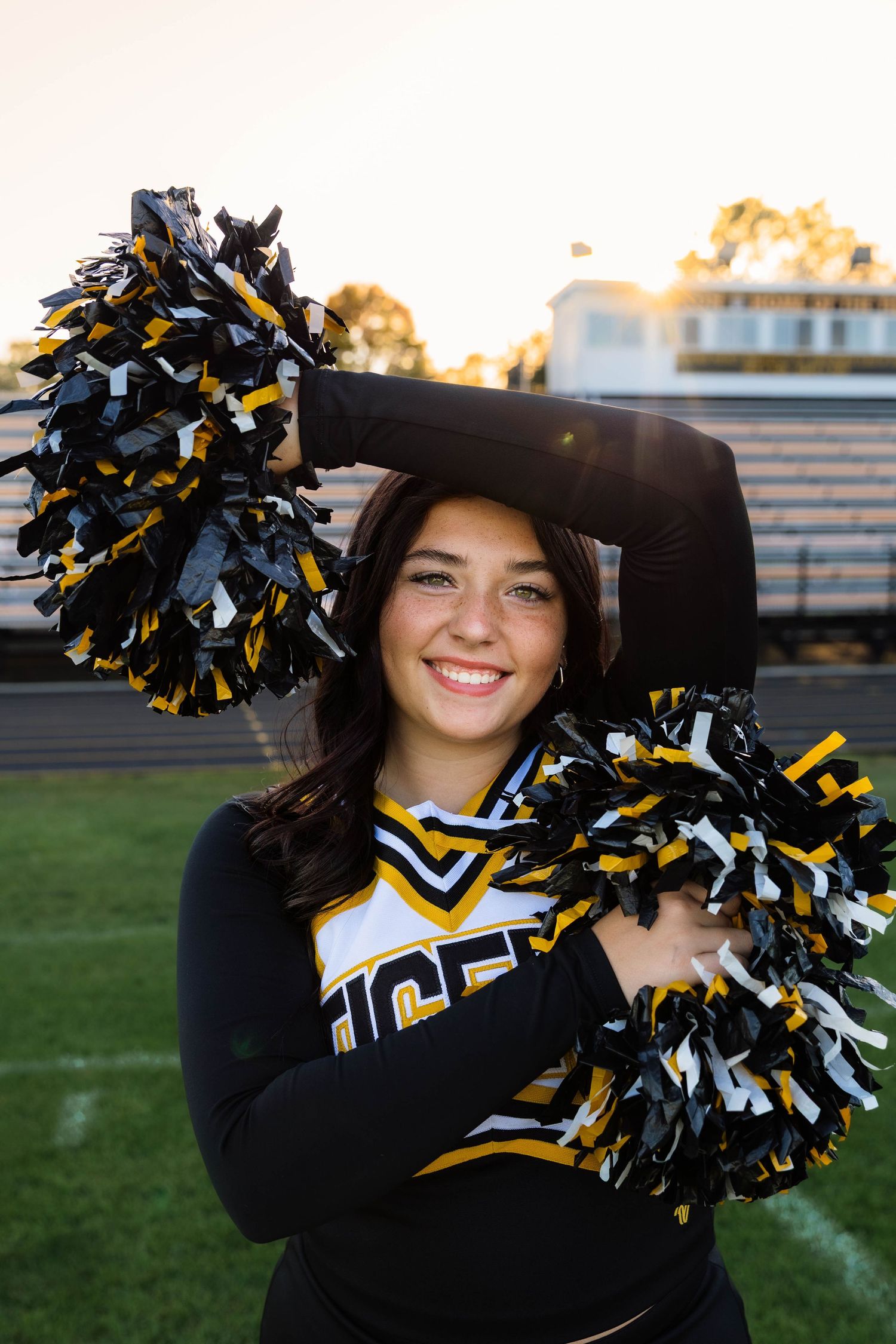 Smiling cheerleader in black and gold Tigers uniform poses with pom-poms on football field at sunset.