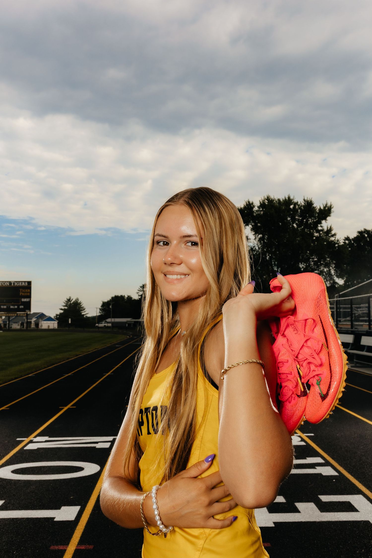 Smiling blonde female track athlete holds pink running spikes on school track at dusk.