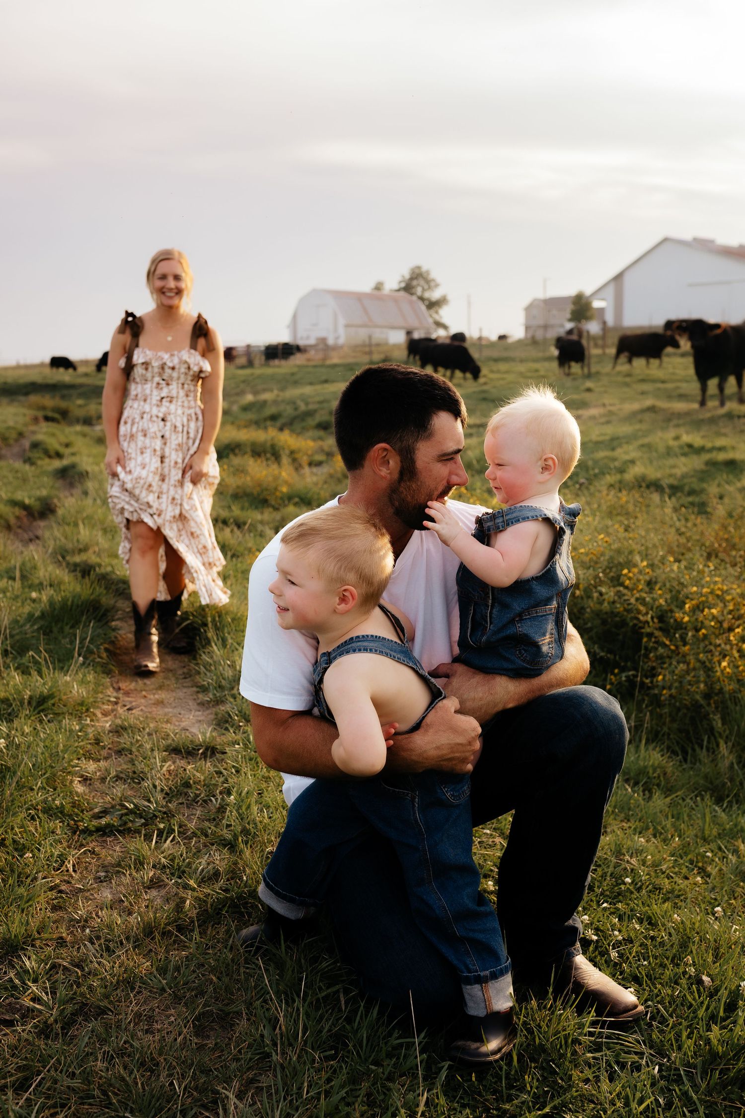 Young family with father holding two toddlers in a green pasture with cattle and barn.