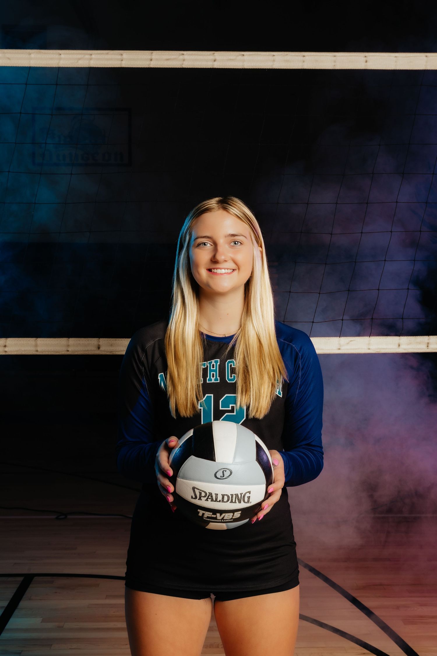 Female volleyball player in uniform holding a Spalding ball in a dramatic smoky gym setting.