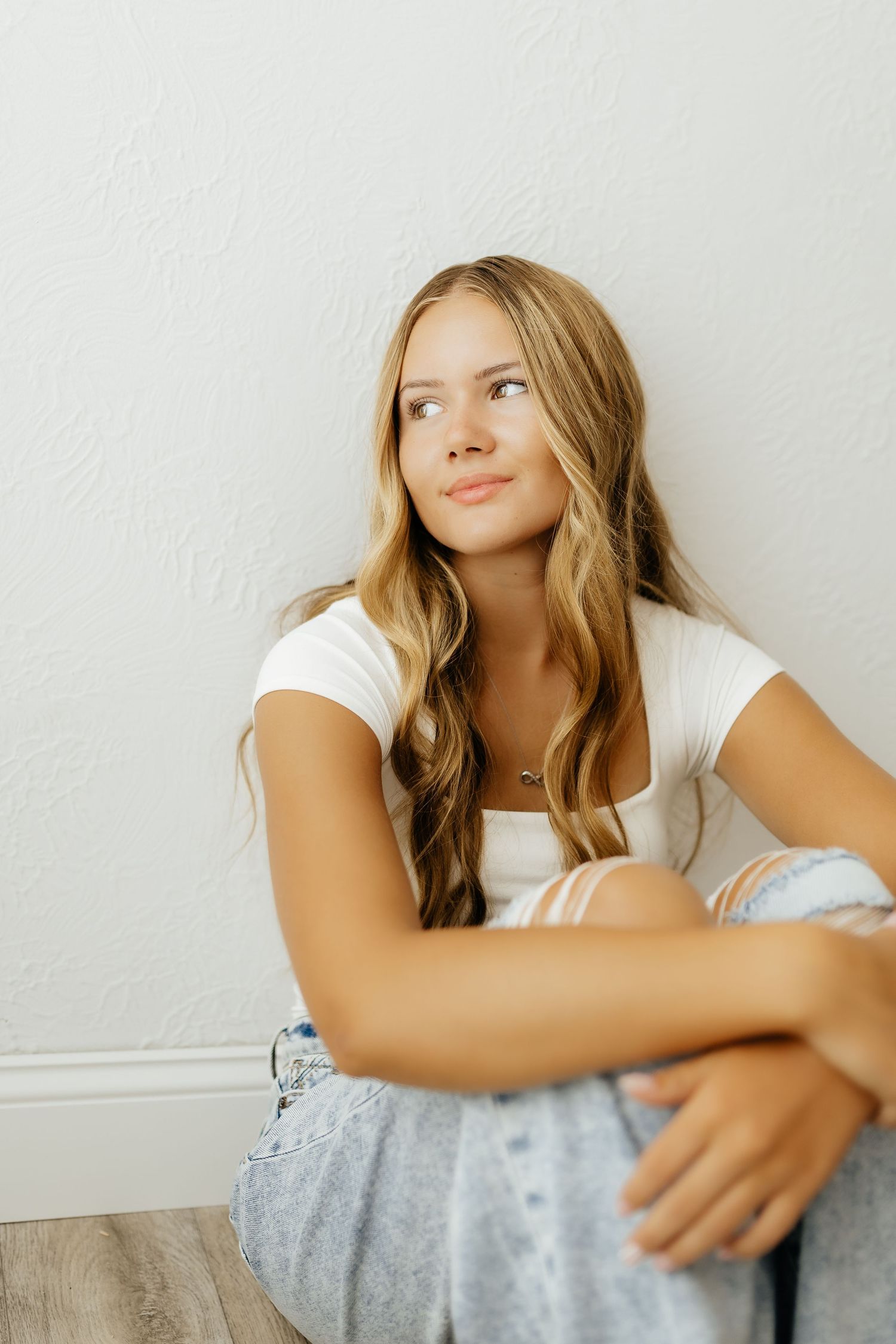 Blonde teenage girl in white top and ripped jeans sits against white wall looking upward.