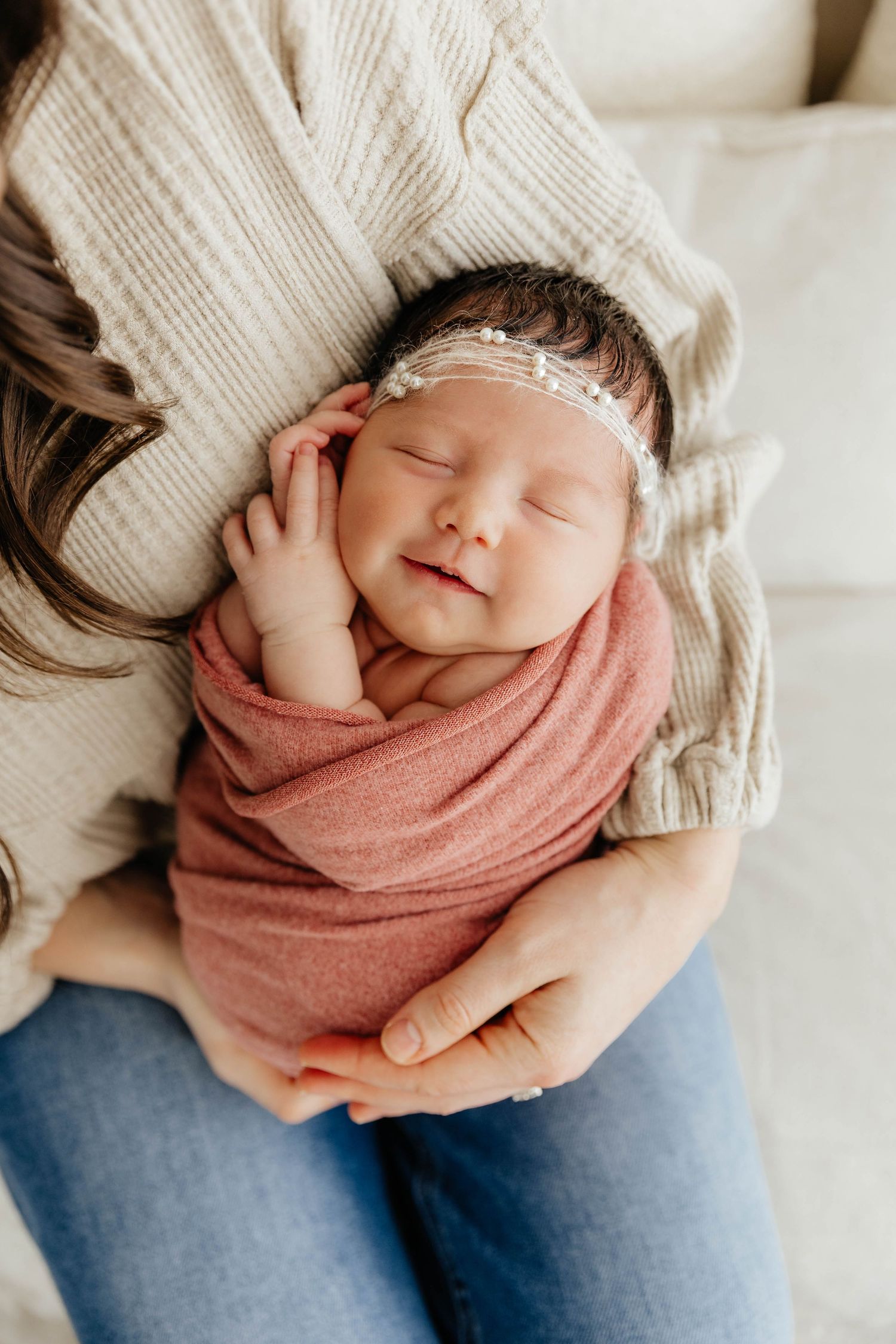 Close-up of sleeping newborn in dusty rose swaddle with pearl headband held against mothers sweater.