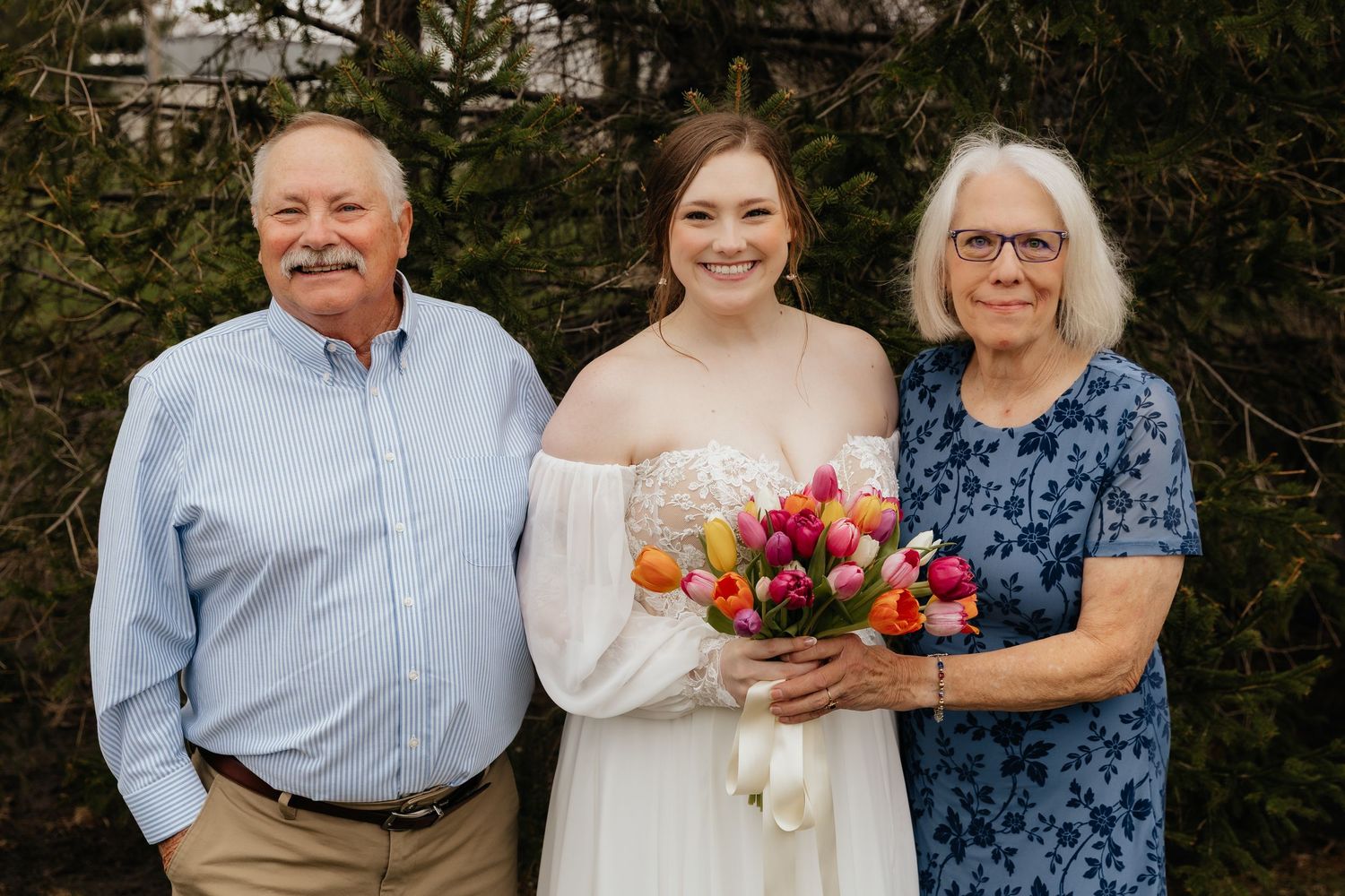 Bride holding colorful tulip bouquet poses with grandparents in outdoor wedding portrait.