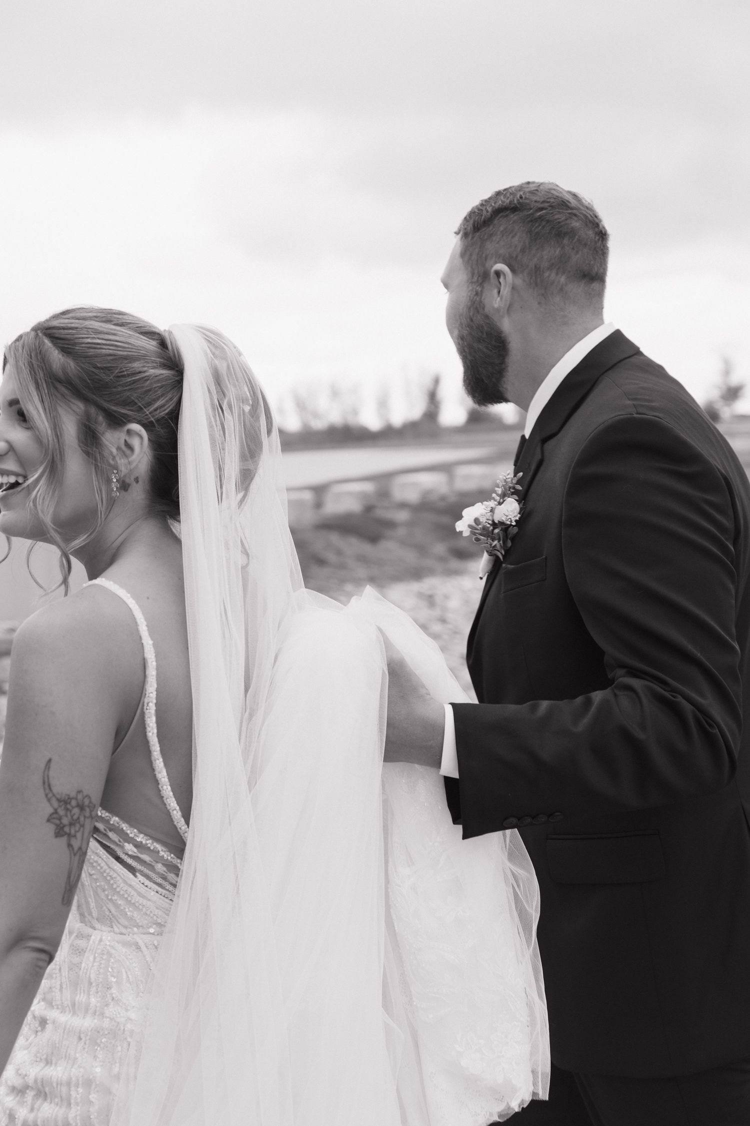Black and white photo of laughing bride and bearded groom walking together outdoors.