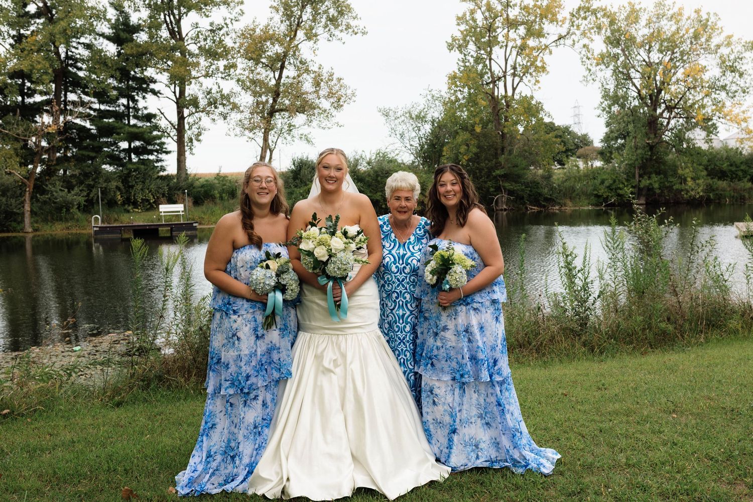 Bride, two bridesmaids, and elderly woman pose together by pond at outdoor wedding.
