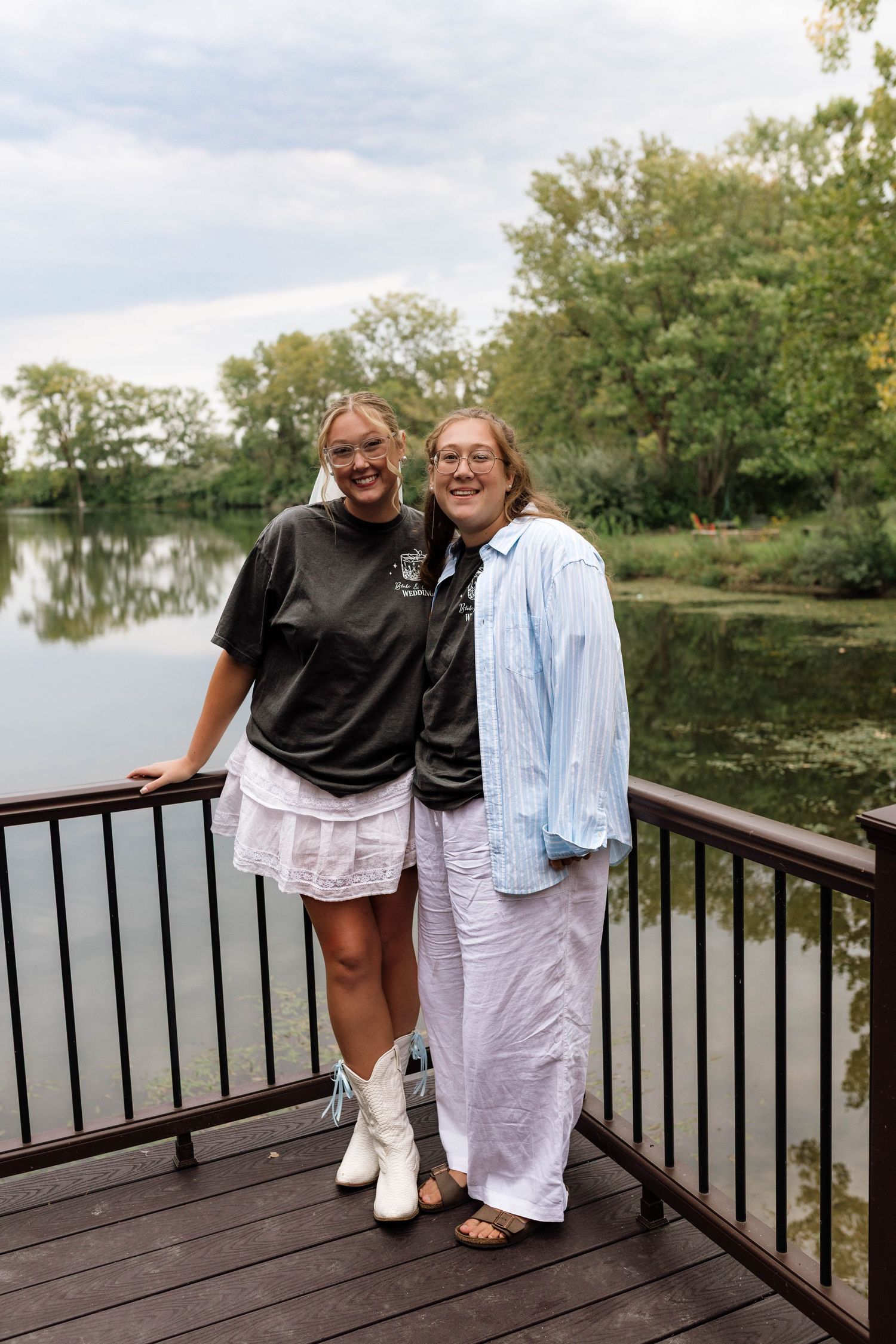 Two young women smiling together on a lakeside deck with lush green trees behind them.