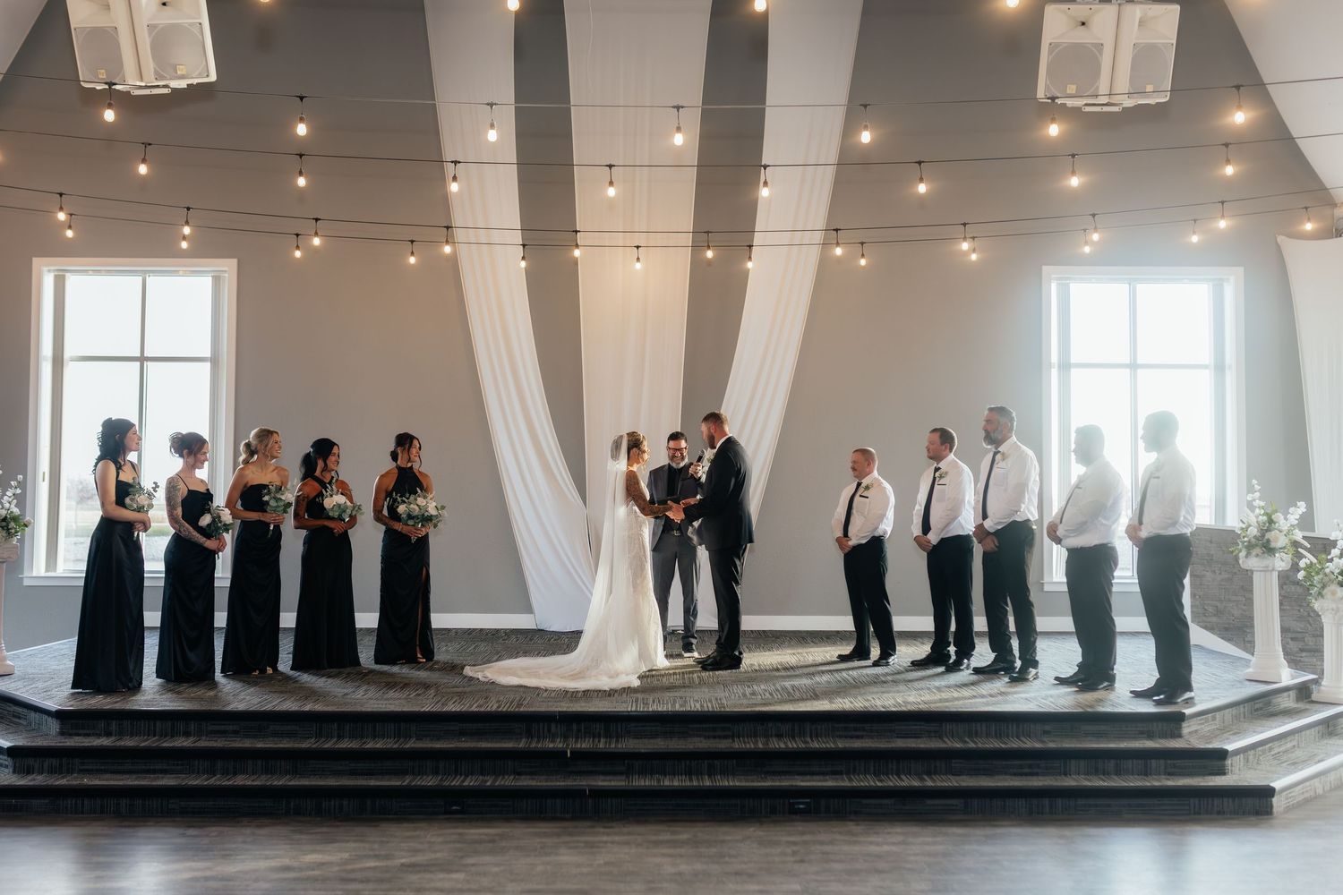 Wedding party stands at altar with string lights and white draping in elegant venue.