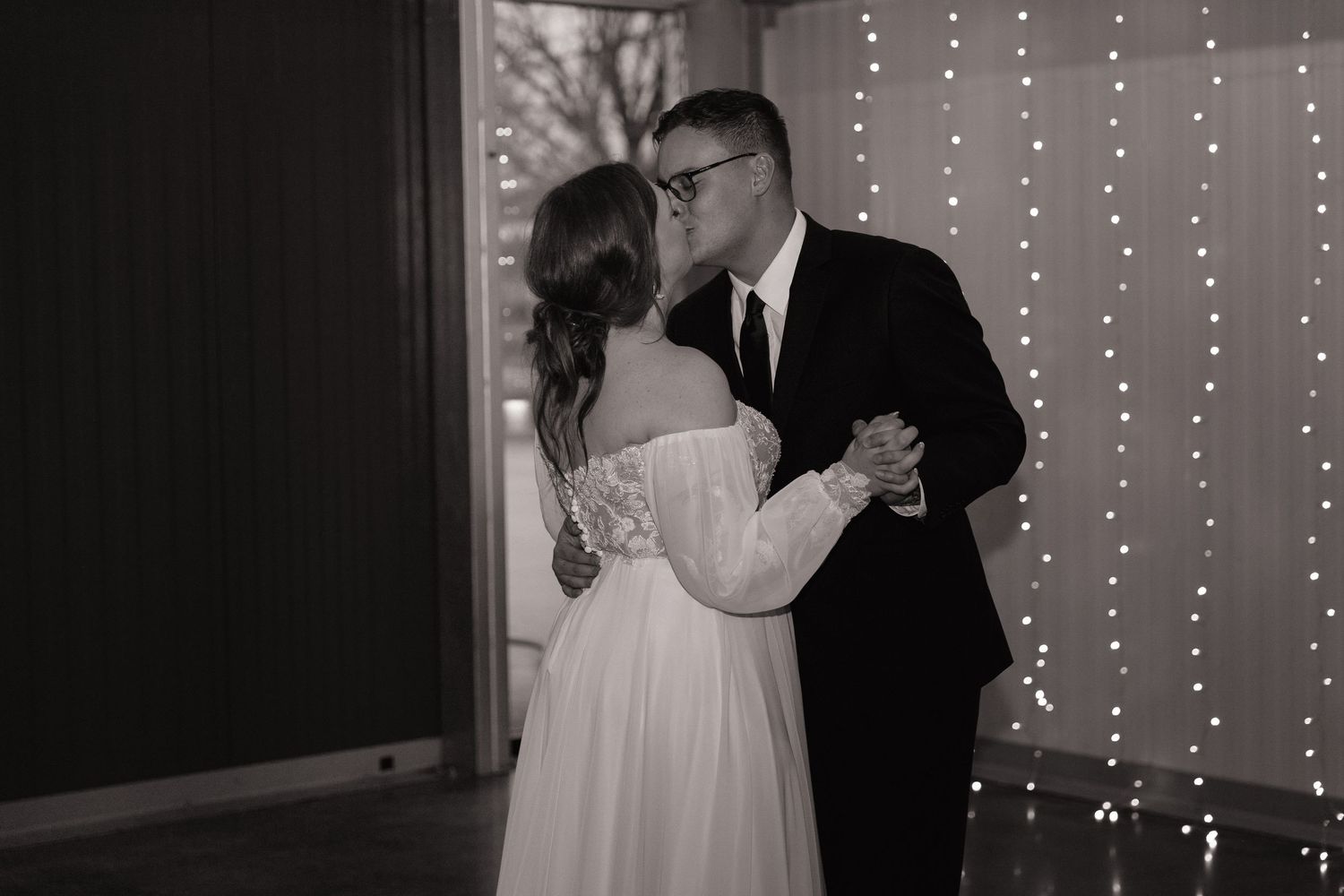 Black and white photo of bride and groom sharing first dance in front of twinkling fairy light backdrop.
