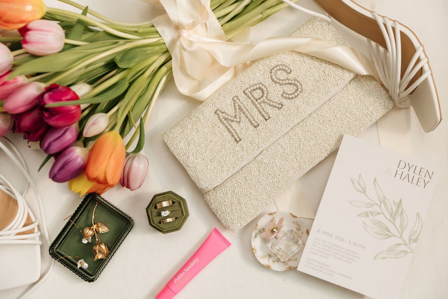 Bridal flat lay with MRS beaded clutch, tulip bouquet, gold earrings, and Dylen Haley wedding invitation.