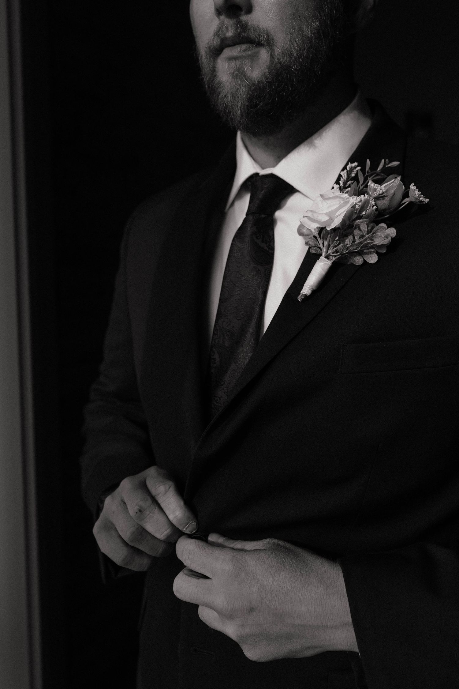 Black and white close-up of a groom in a dark suit buttoning his jacket with a floral boutonniere.