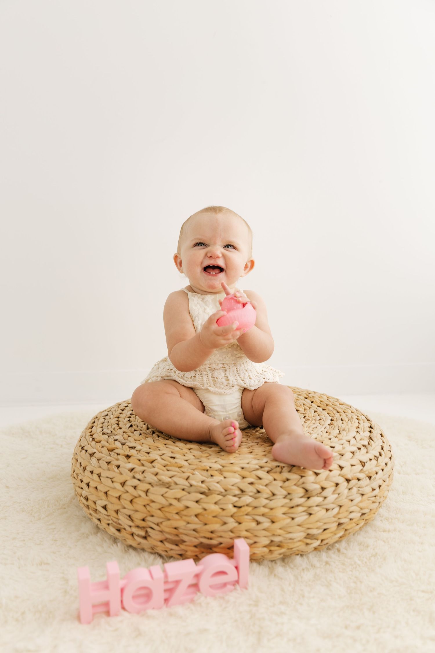 Laughing baby girl named Hazel sitting on woven pouf holding pink toy with name sign on floor.
