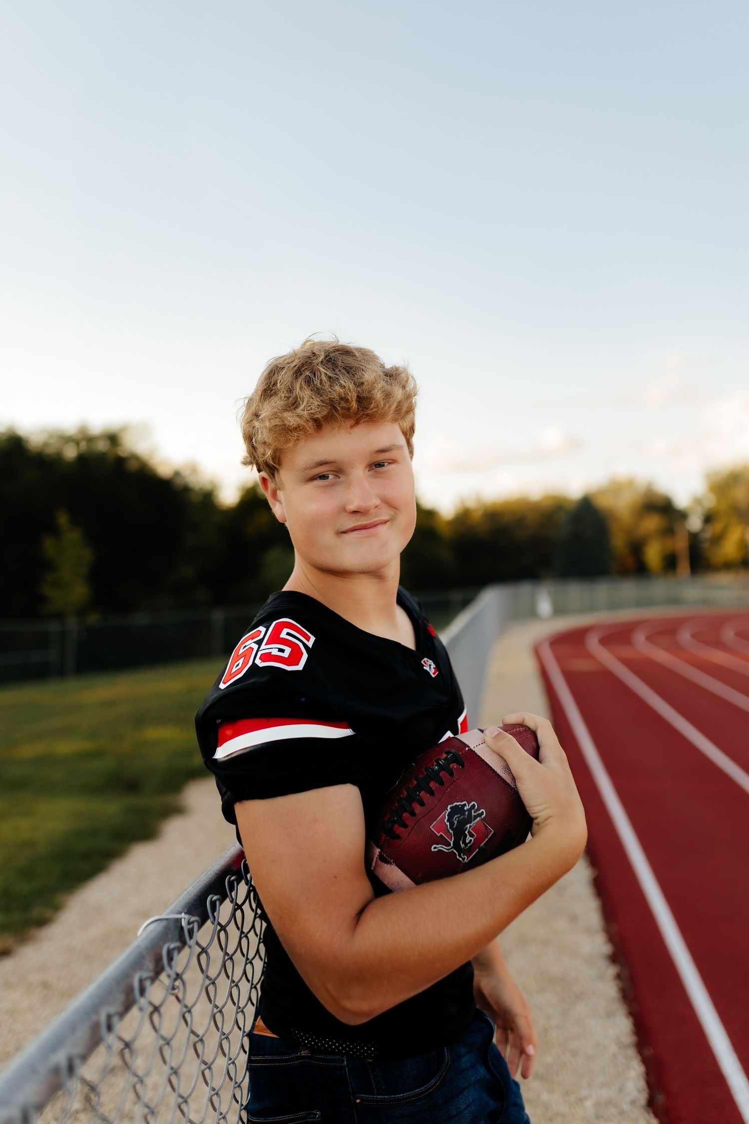 Smiling young football player in black number 66 jersey holding ball by a track fence.