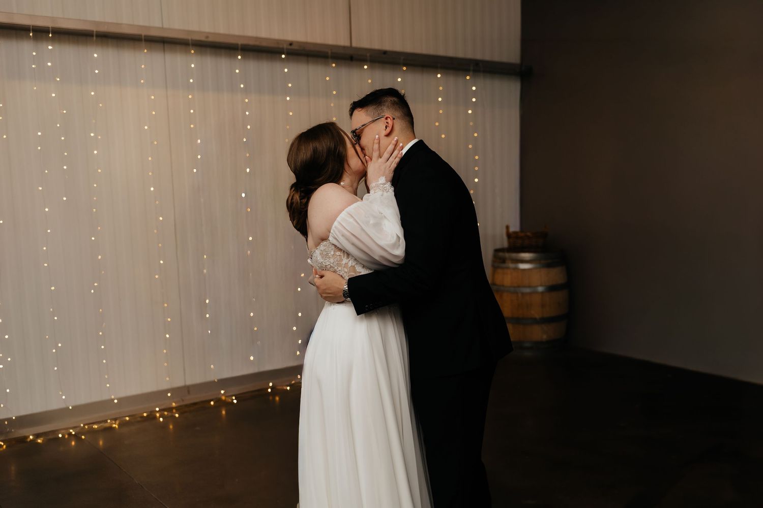 Newlyweds share first kiss on dance floor in front of twinkling fairy light backdrop.