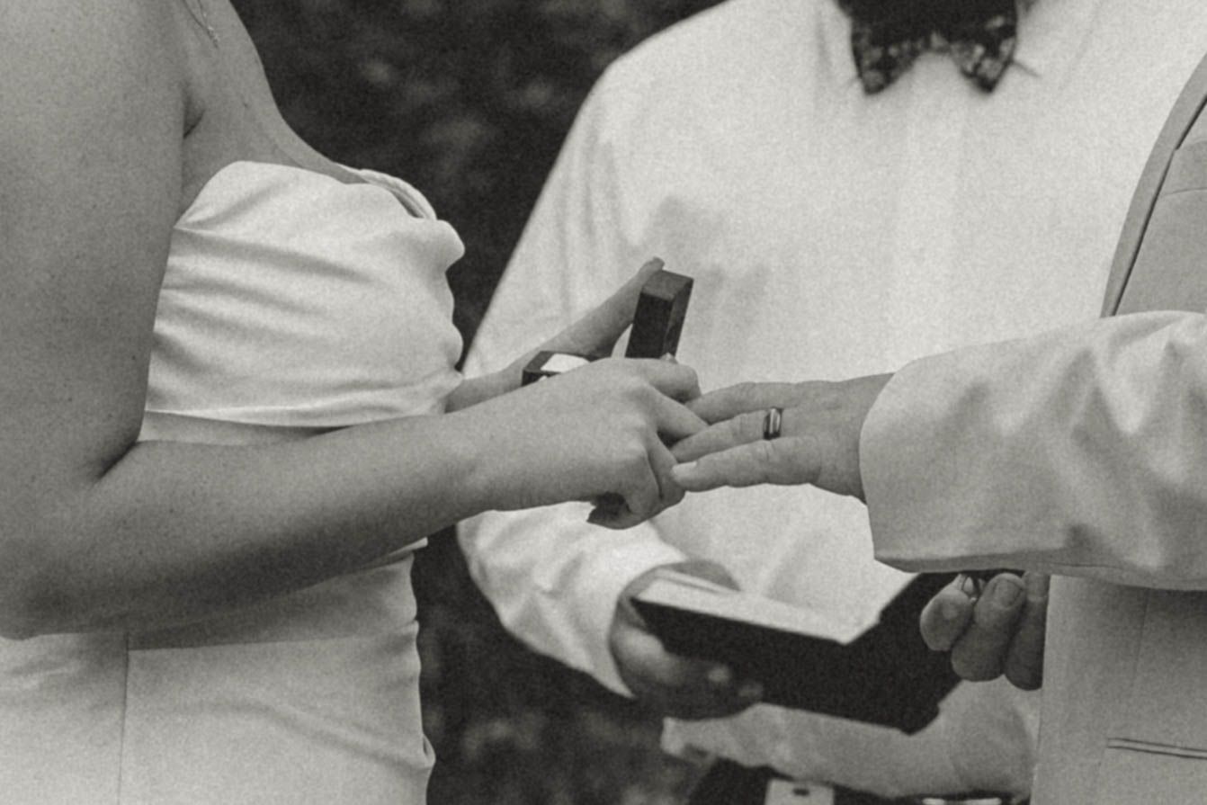 Black and white photo of couple exchanging wedding rings during outdoor ceremony.