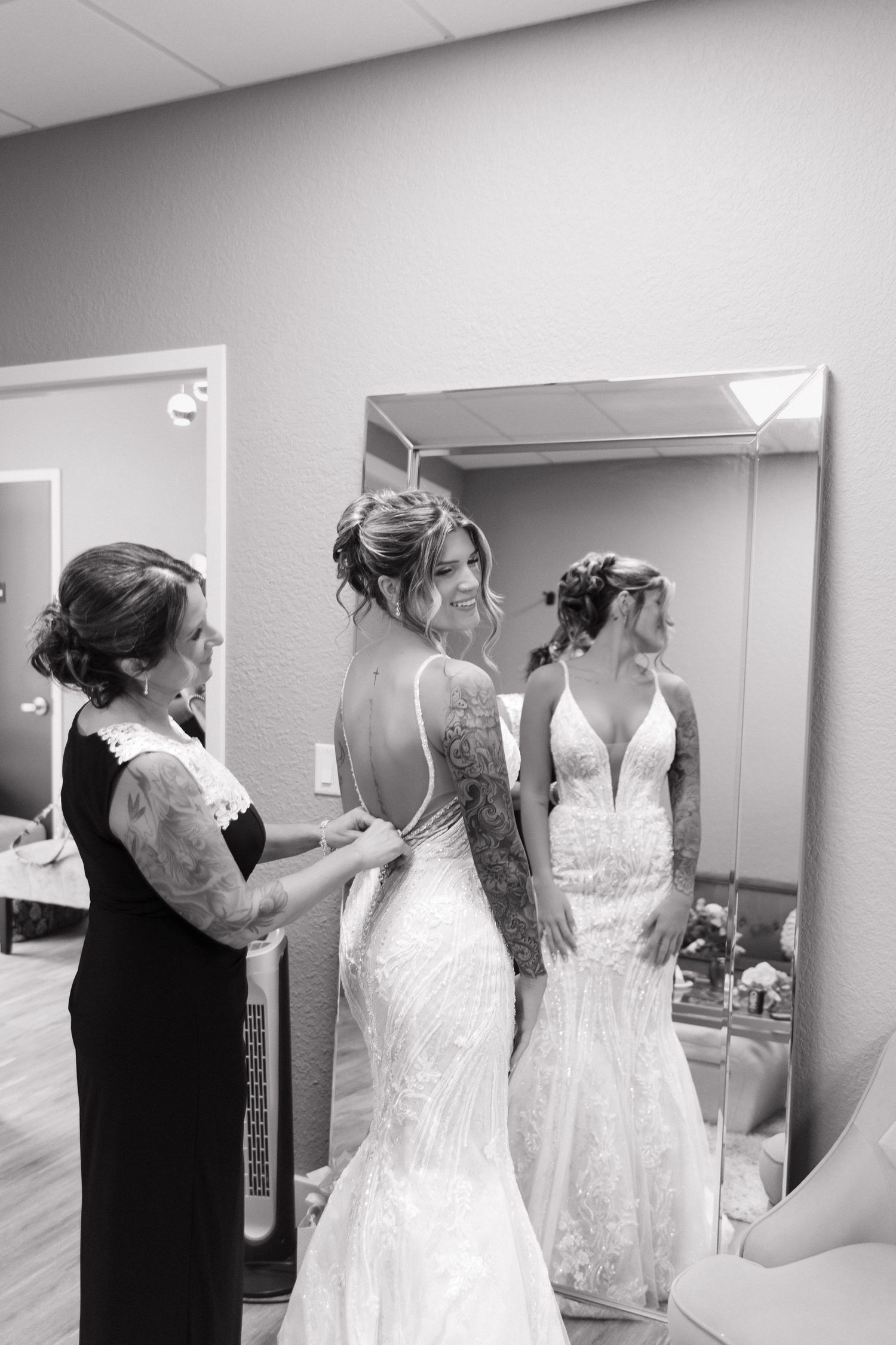 Black and white photo of mother helping tattooed bride button her lace wedding gown.